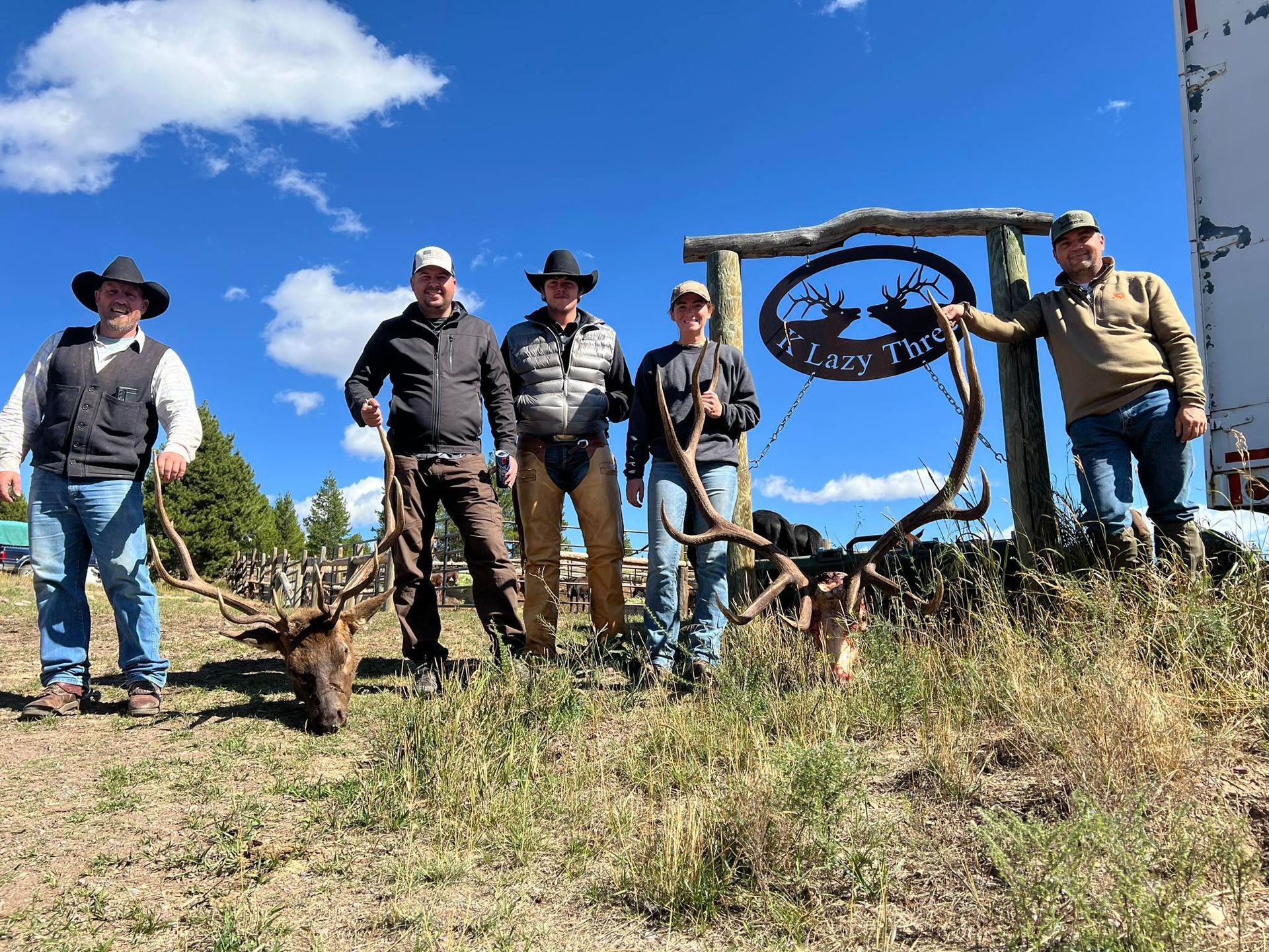 A group of people standing in from of a K Lazy Three sign and posing with a couple mule deer heads