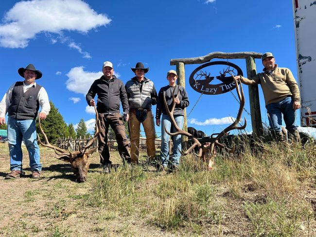 A group of people standing in from of a K Lazy Three sign and posing with a couple mule deer heads