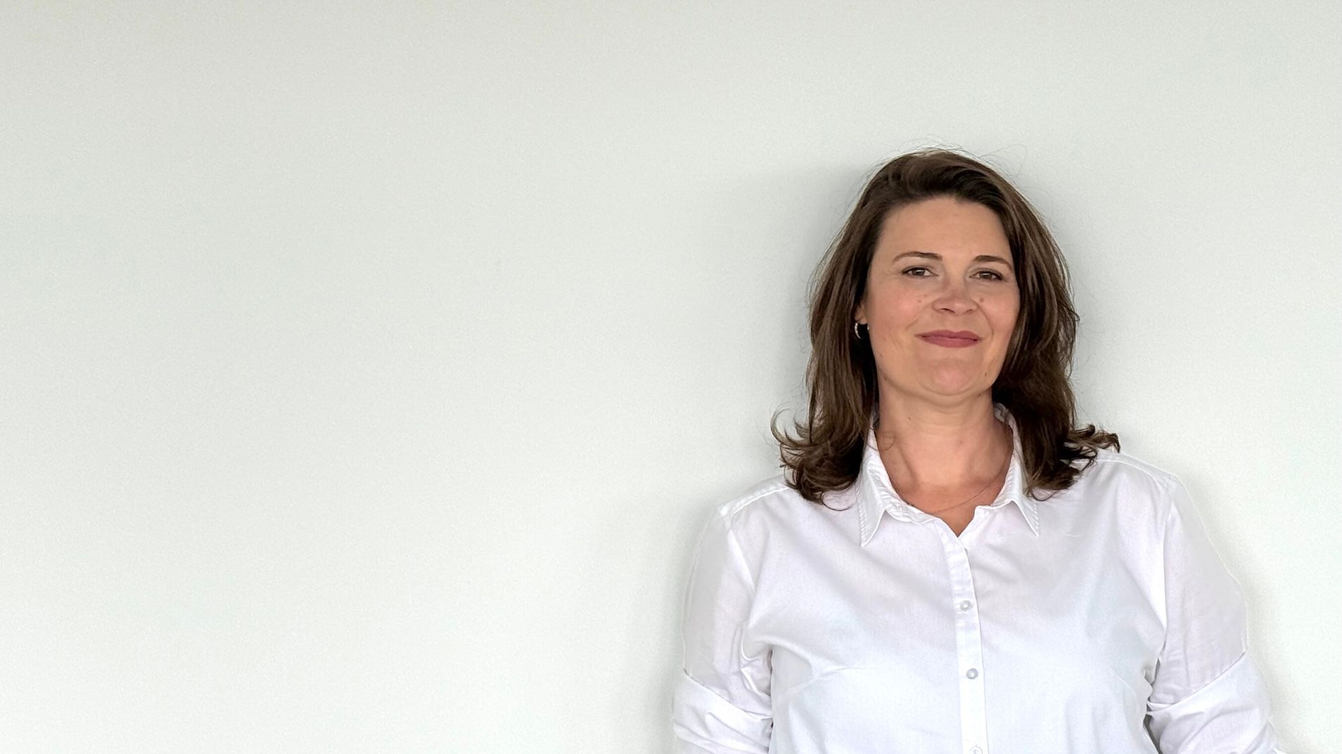 Eine Frau in einem weißen Hemd steht vor einer weißen Wand.