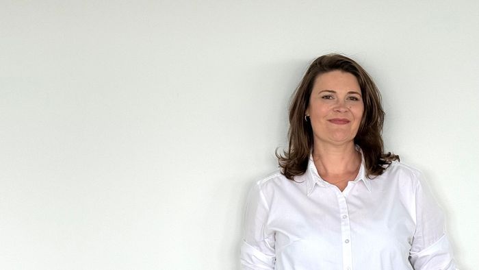 Eine Frau in einem weißen Hemd steht vor einer weißen Wand.