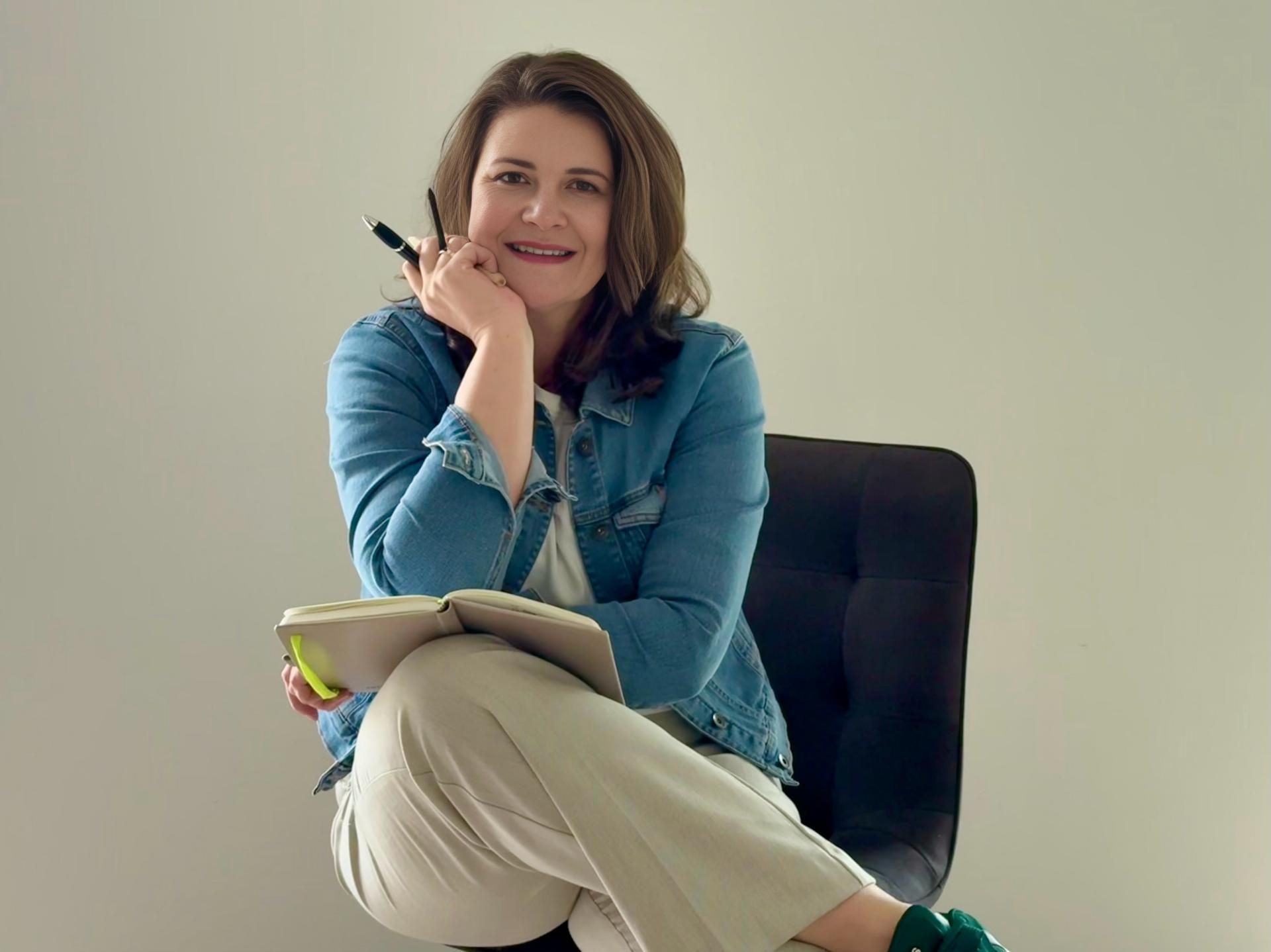 Eine Frau sitzt auf einem Stuhl und hält einen Stift und ein Notizbuch in der Hand.