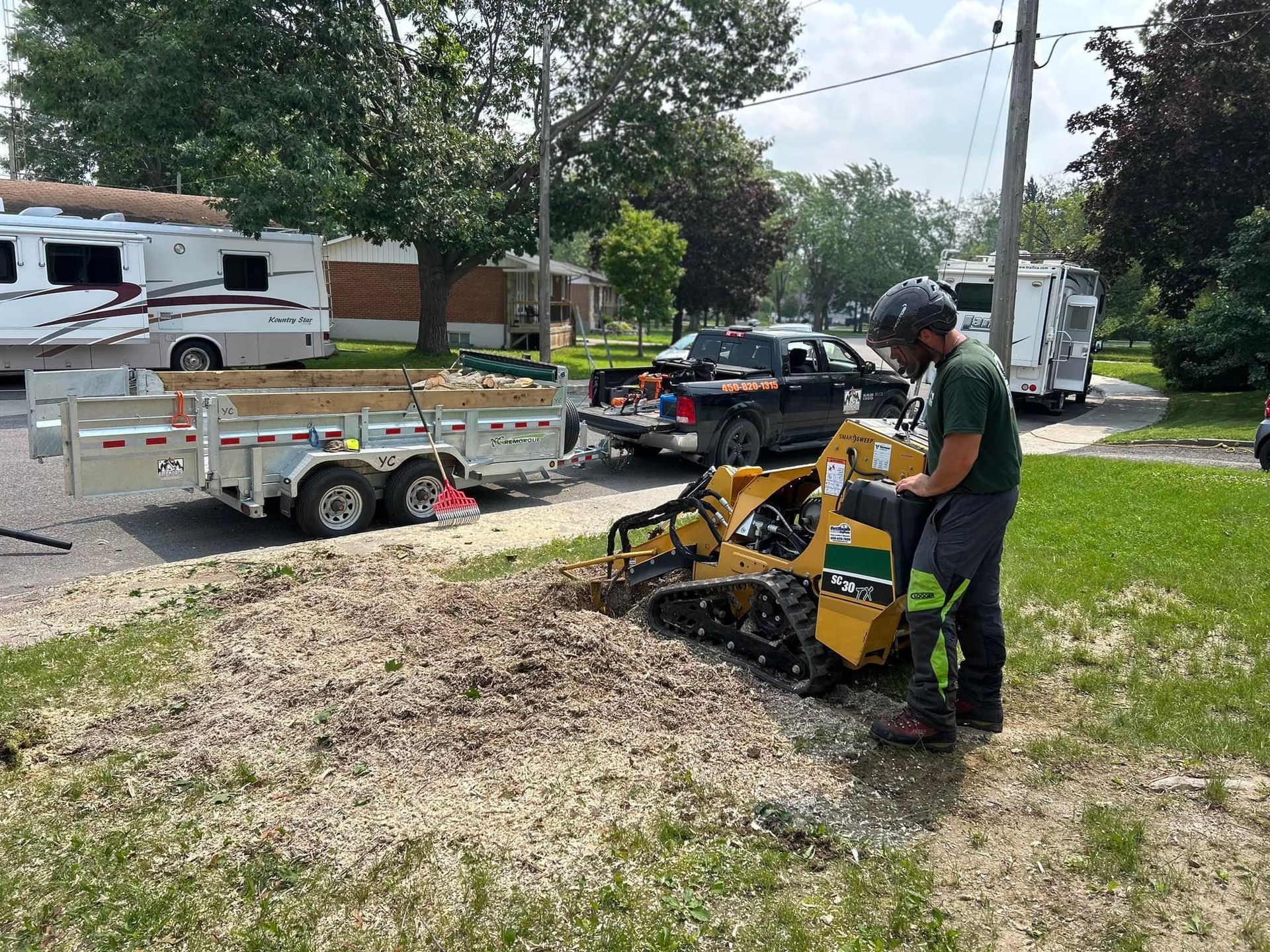 Une personne utilise une dessoucheuse dans une zone résidentielle ; une remorque et des caravanes sont visibles.