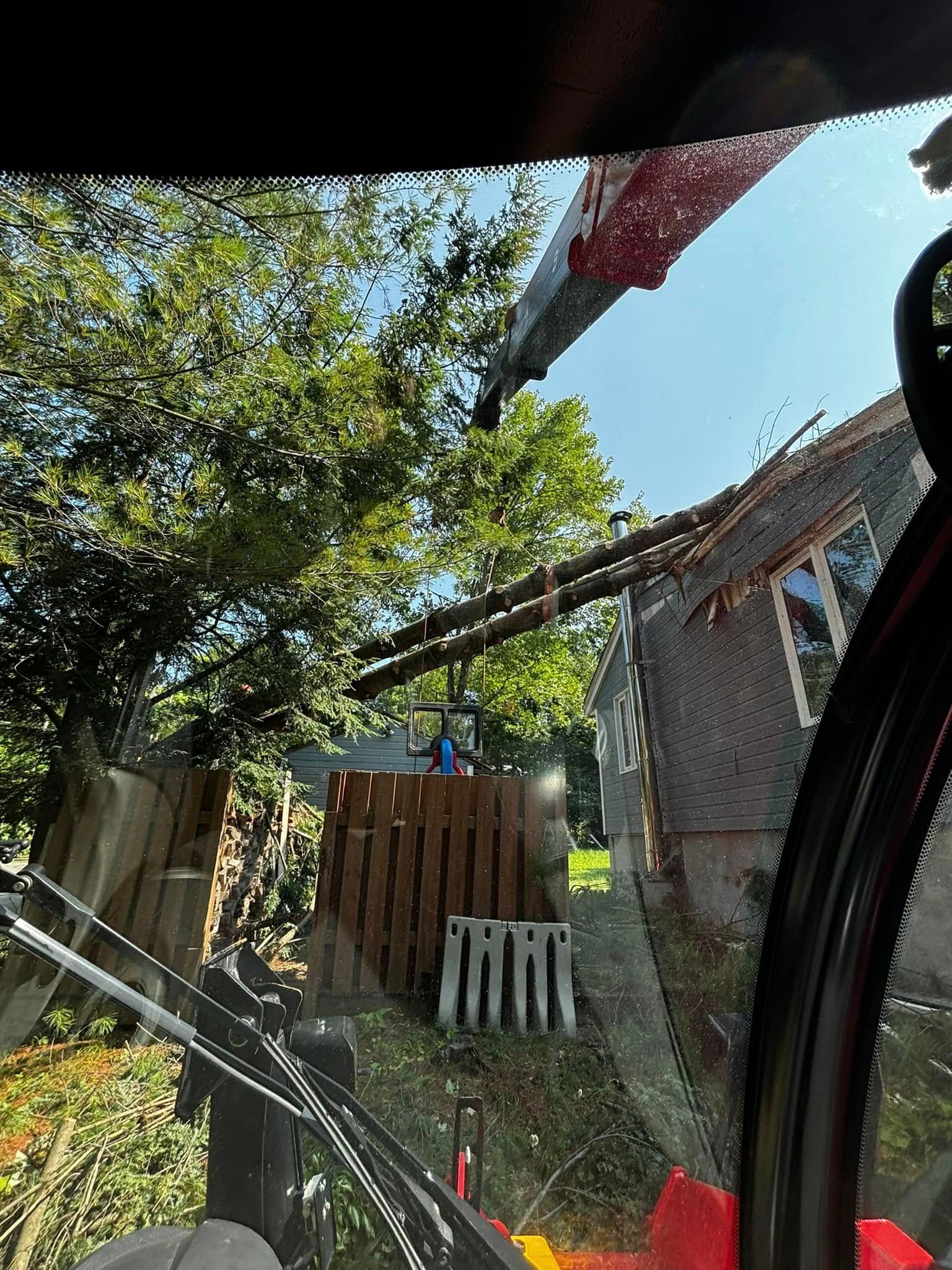 Vue depuis la cabine d'un engin de chantier : une toiture est en cours de dépose. Une poutre en bois surplombe un bâtiment. Arbres et clôture se trouvent à l'arrière-plan.