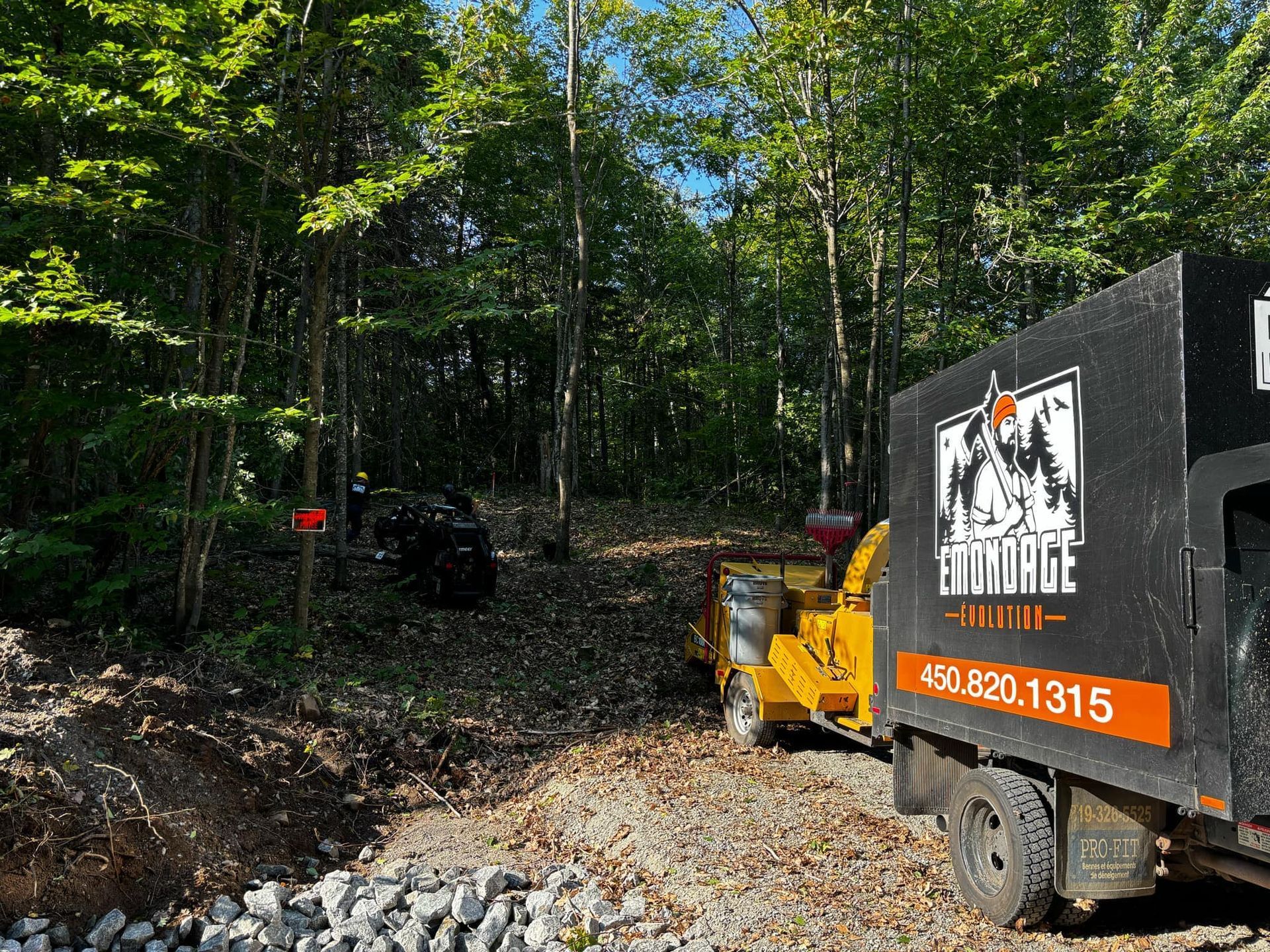 Un camion équipé d'une déchiqueteuse à bois traite des débris de bois dans une zone boisée ; une voiture est visible en arrière-plan.