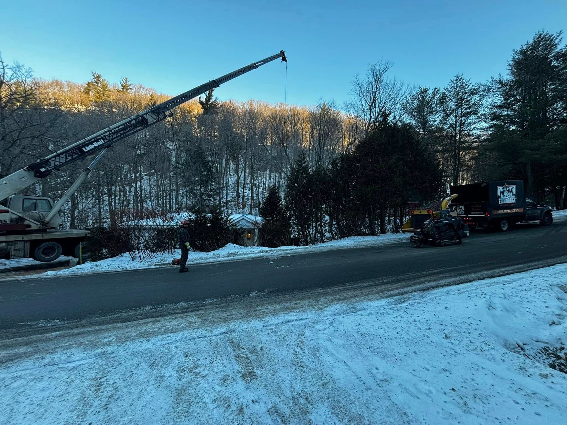 Une grue soulève quelque chose près d'une route enneigée, avec une personne dans la rue et des arbres en arrière-plan.