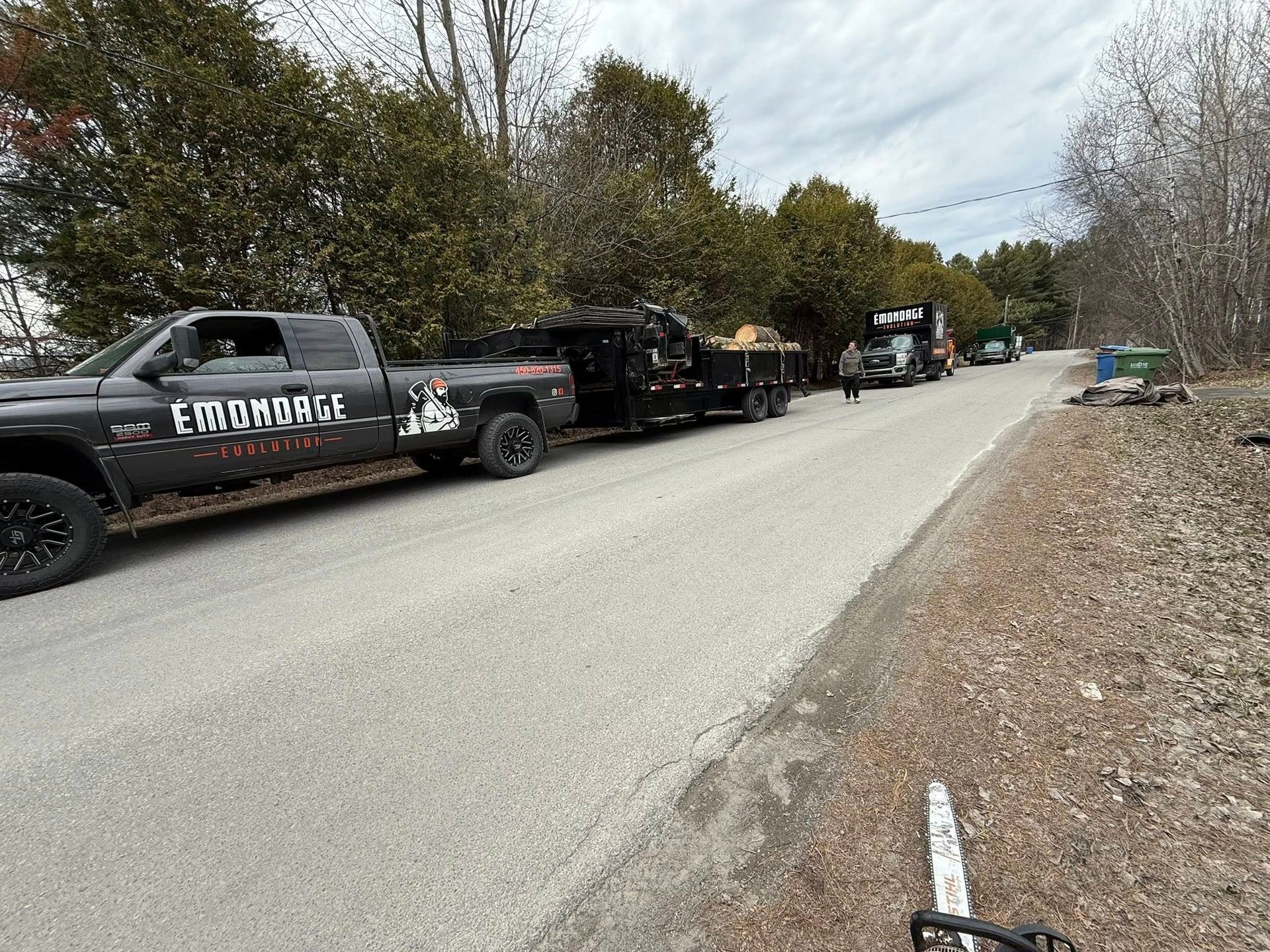 Camion avec remorque sur une route, logo d'une entreprise d'élagage, personne marchant vers un camion, tronçonneuse au premier plan.