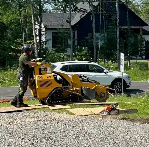 Un homme utilise une dessoucheuse jaune sur une propriété résidentielle. Un SUV blanc est stationné à proximité.