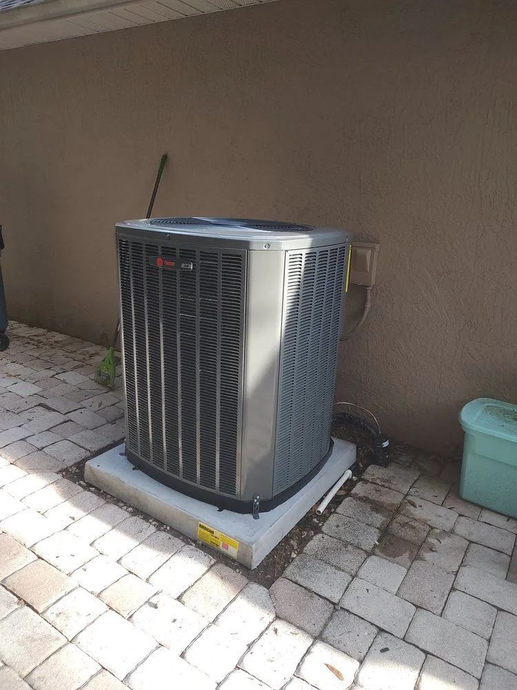 A dark grey Trane air conditioning unit sits on a concrete pad atop a paver patio next to an exterior house wall.