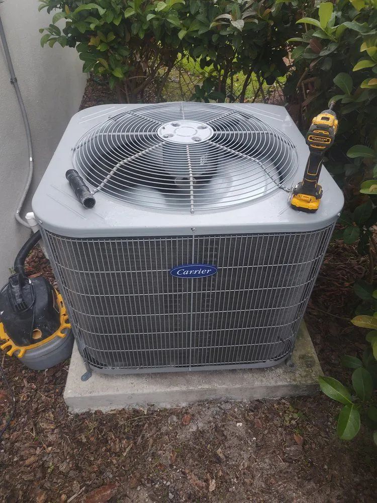 A Carrier air conditioning unit with a drill resting on top, in an outdoor setting with vegetation.