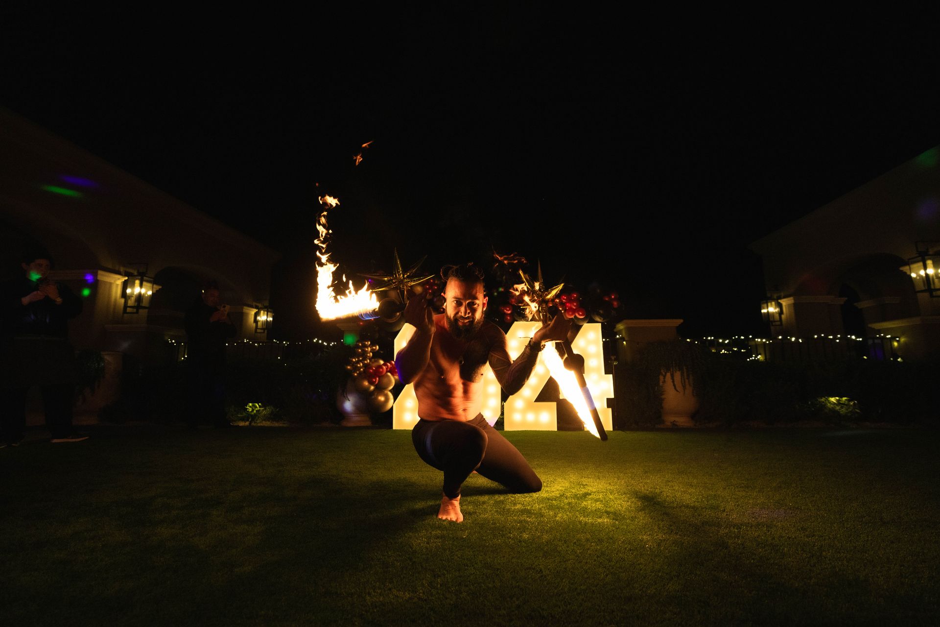 A man is kneeling down in the grass holding a fire stick.