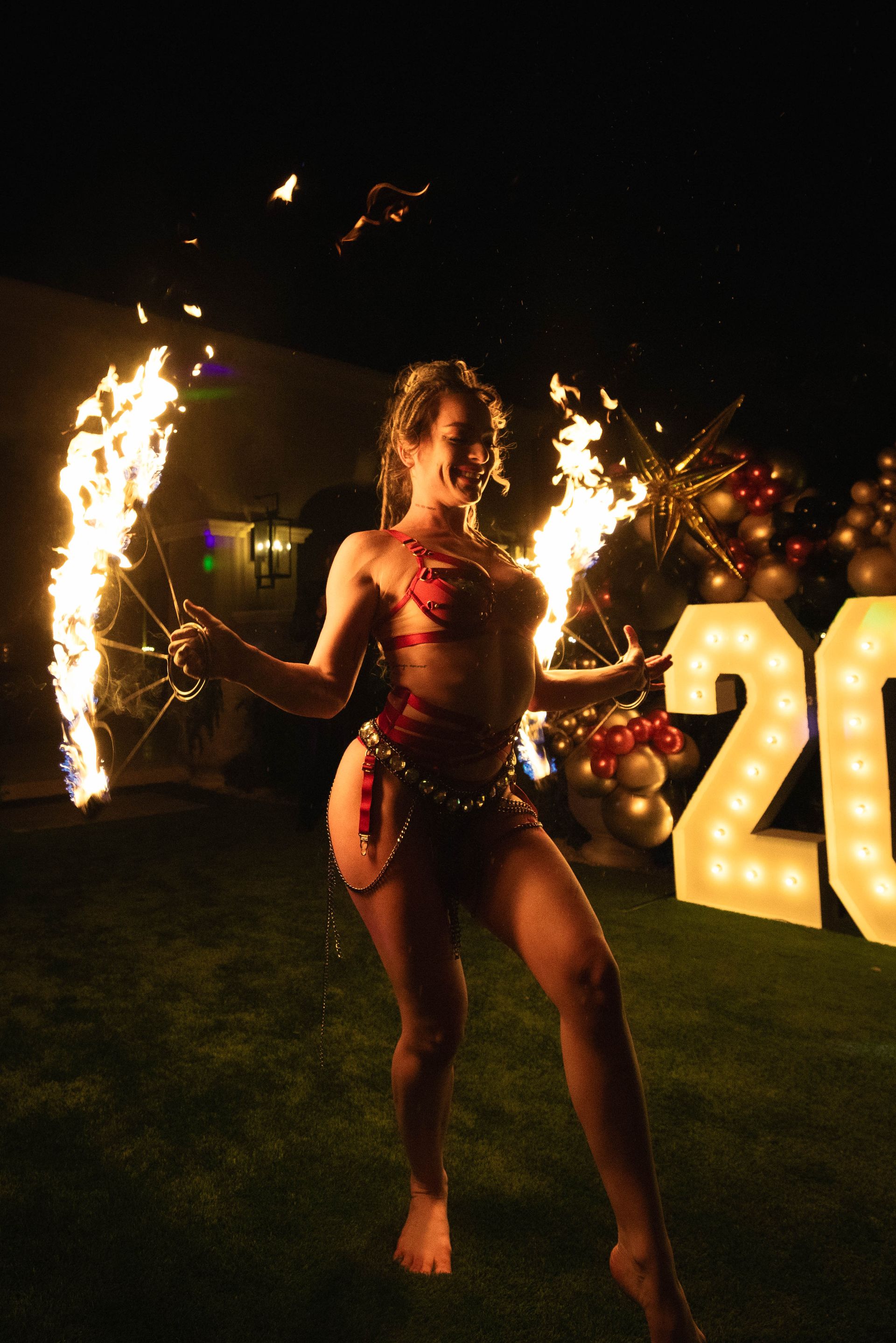 A woman in a bikini is holding two fire torches in front of a sign that says 20.