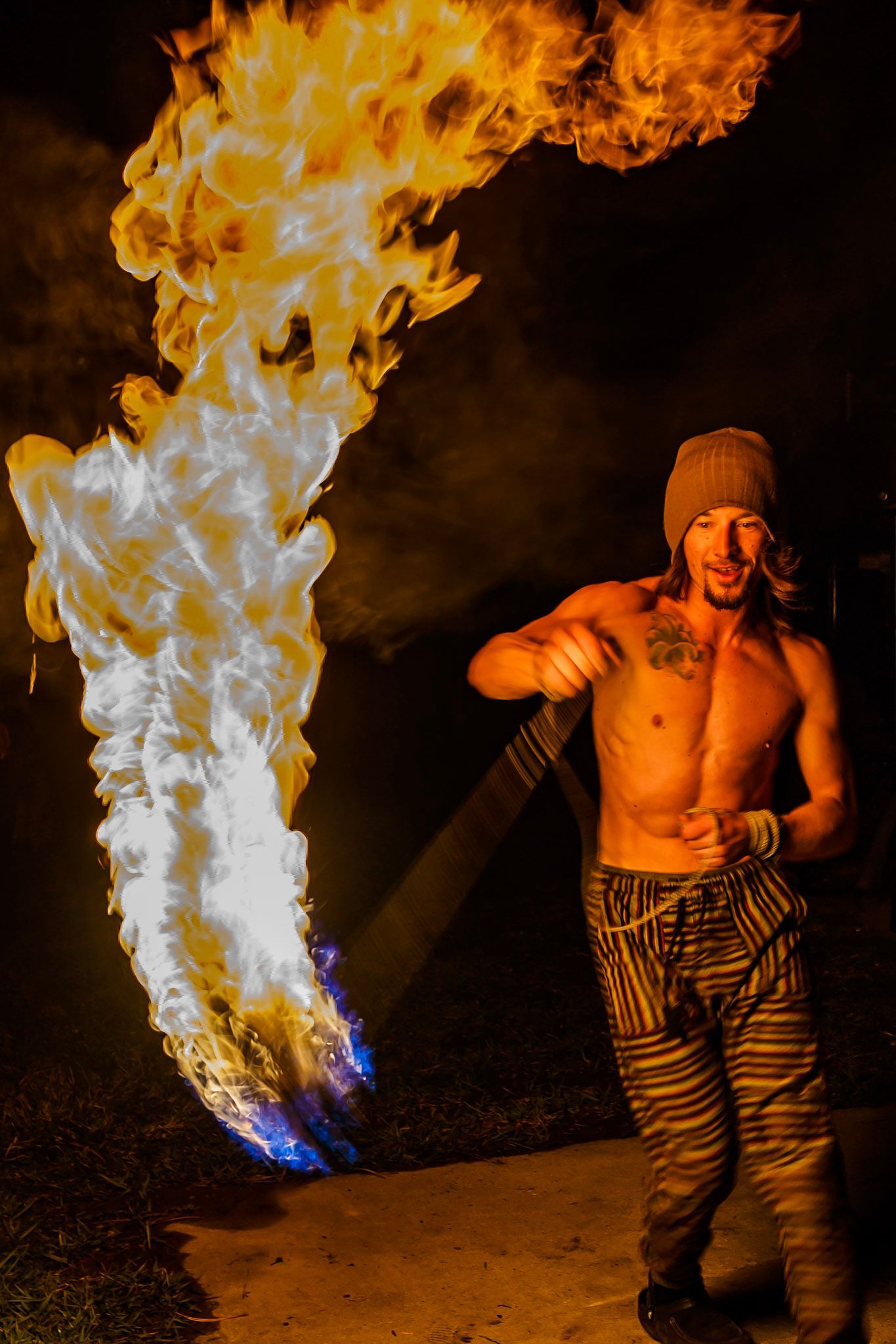 A shirtless man is standing in front of a large flame coming out of his mouth.