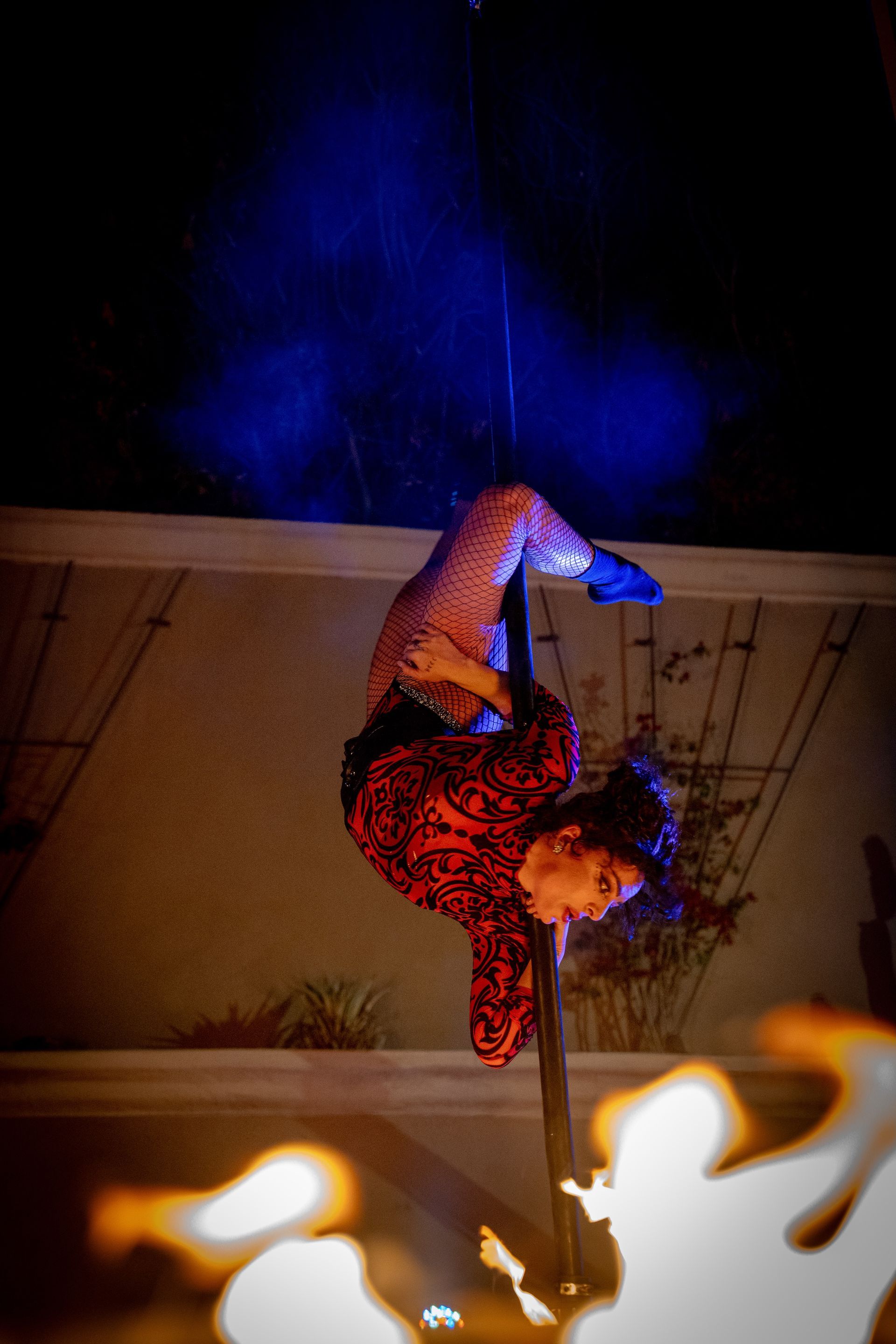 A woman is doing aerial acrobatics on a pole in front of a fire.