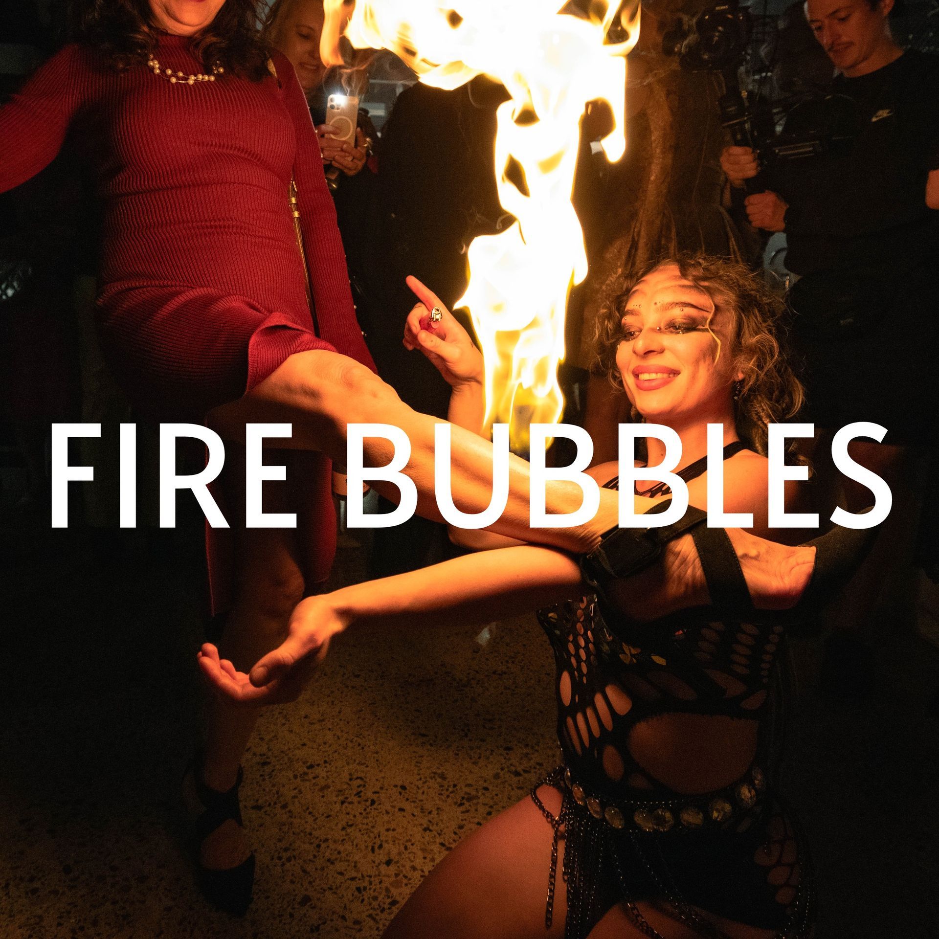 Woman kneels near flames, another in red dress dances. Fire bubbles text on image. Nighttime setting.