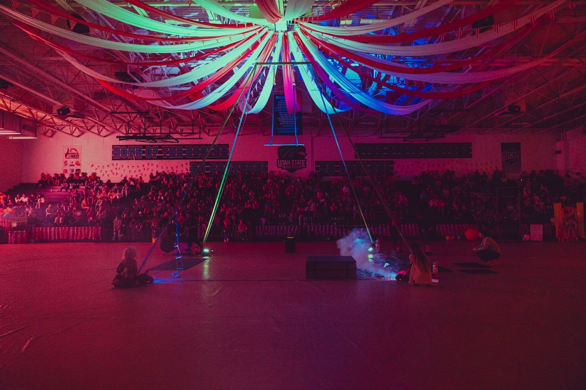 A large crowd of people are sitting in a gym with a colorful ceiling decoration.