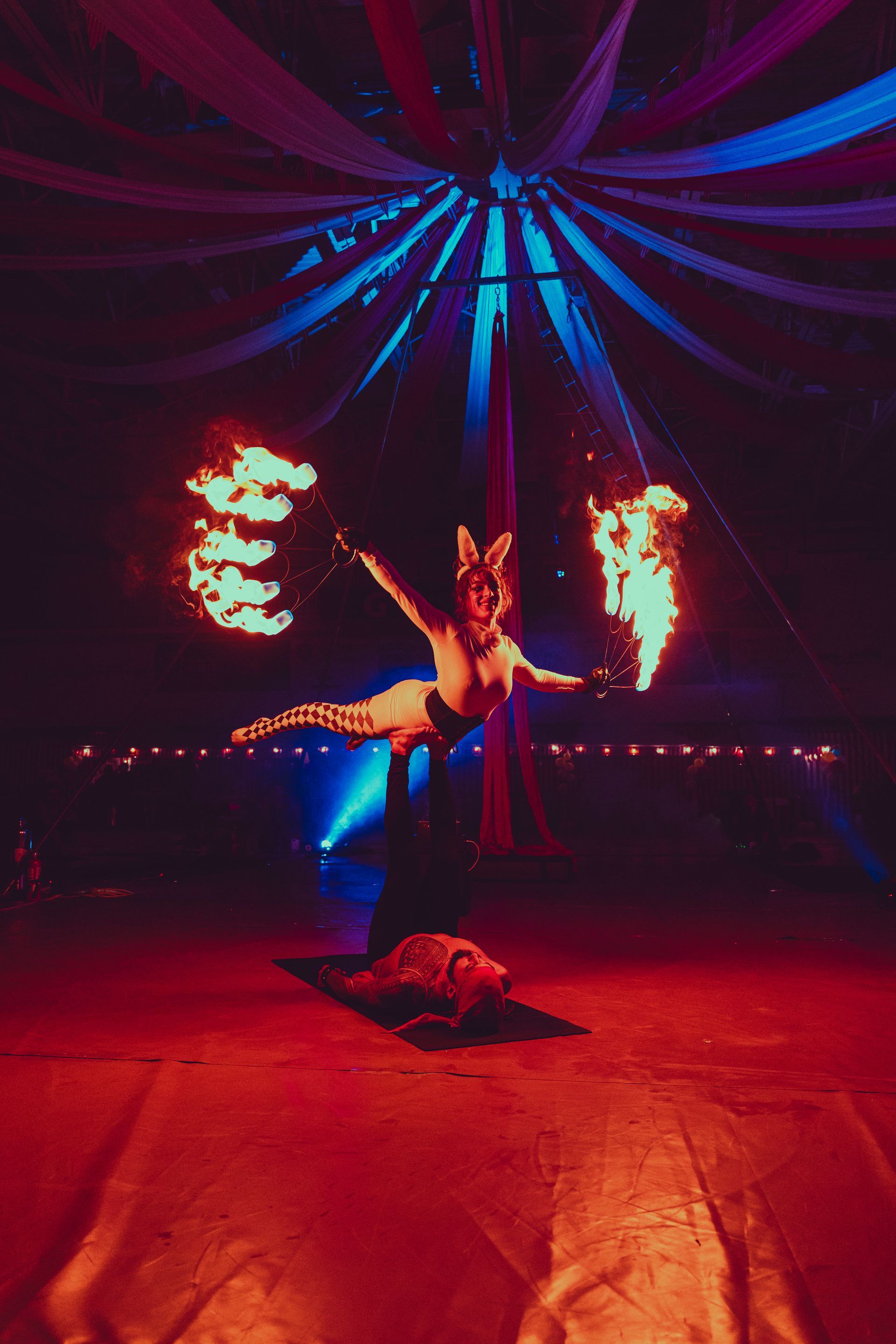 A woman is performing aerial acrobatics in a circus tent with fire coming out of the ceiling.