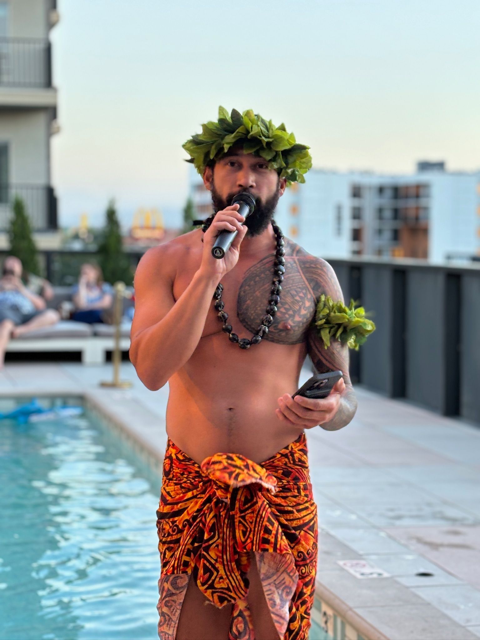 A shirtless man is standing next to a pool holding a microphone.