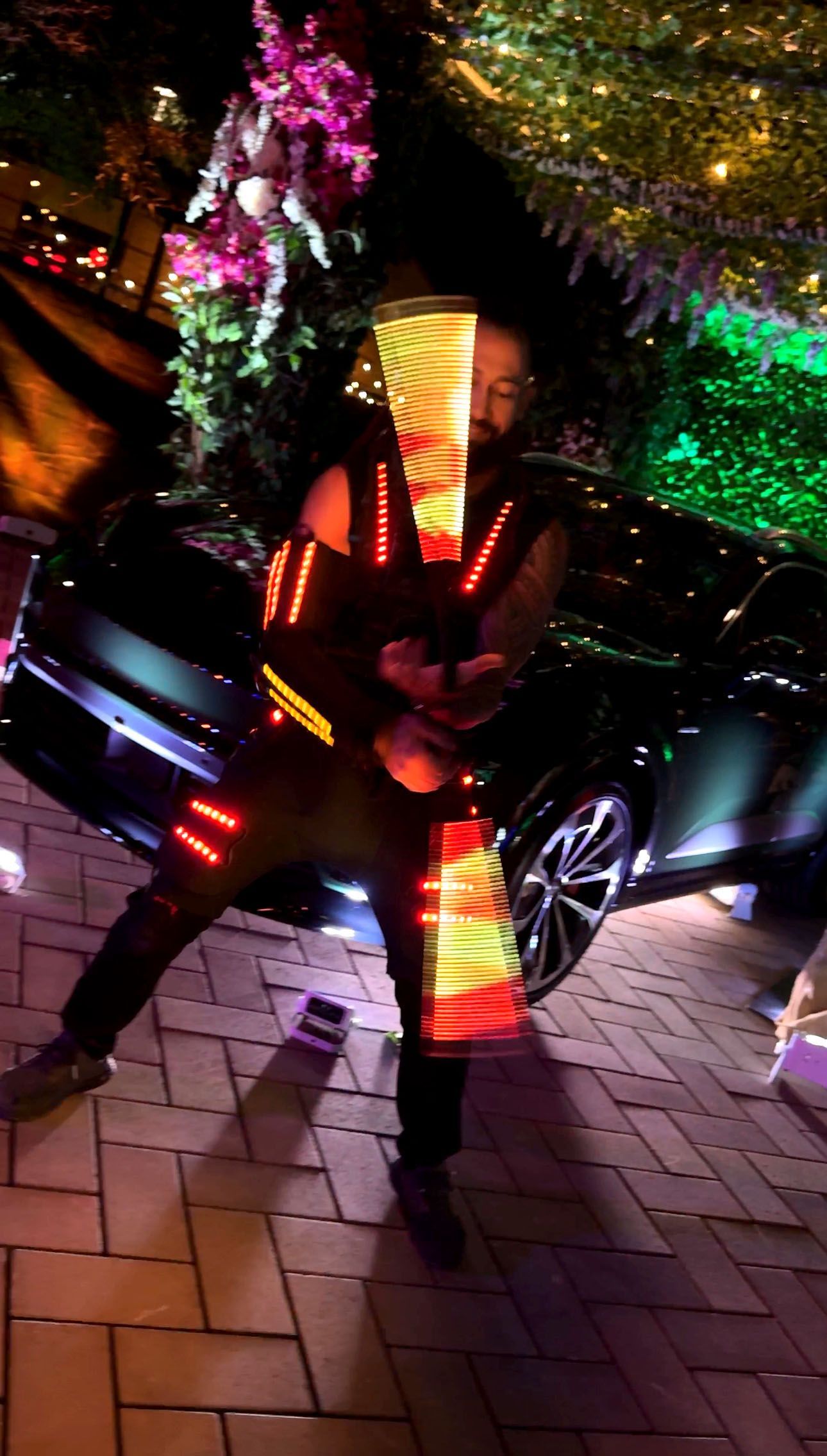 A man in a costume with lights on his arms is standing in front of a car.