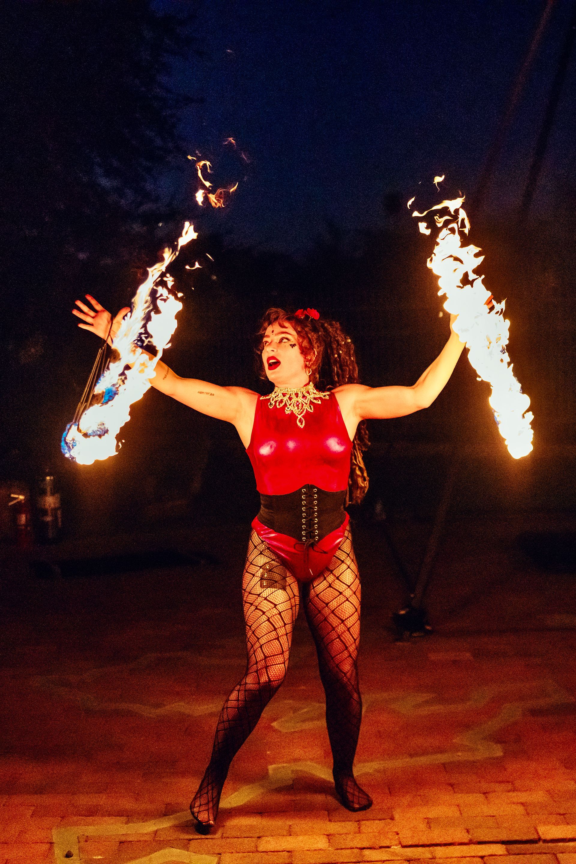 A woman in a red top and black tights is holding two flaming swords.