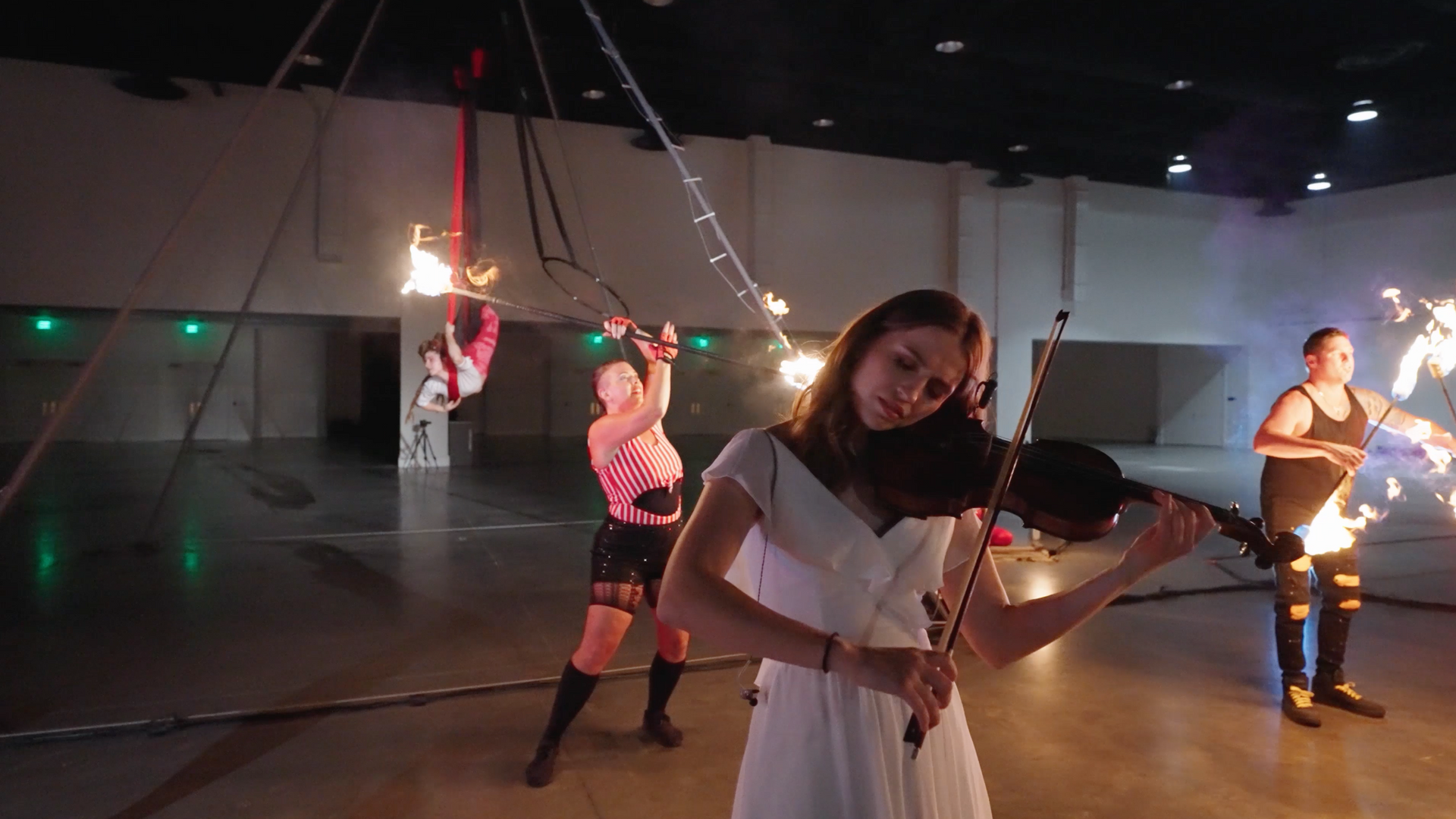 A woman in a white dress is playing a violin in front of a group of fire performers.