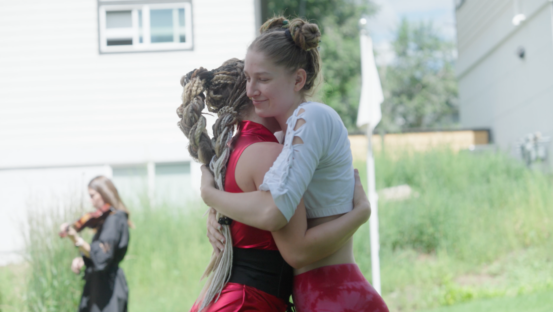 Two women are hugging each other in a field with a woman playing a violin in the background.