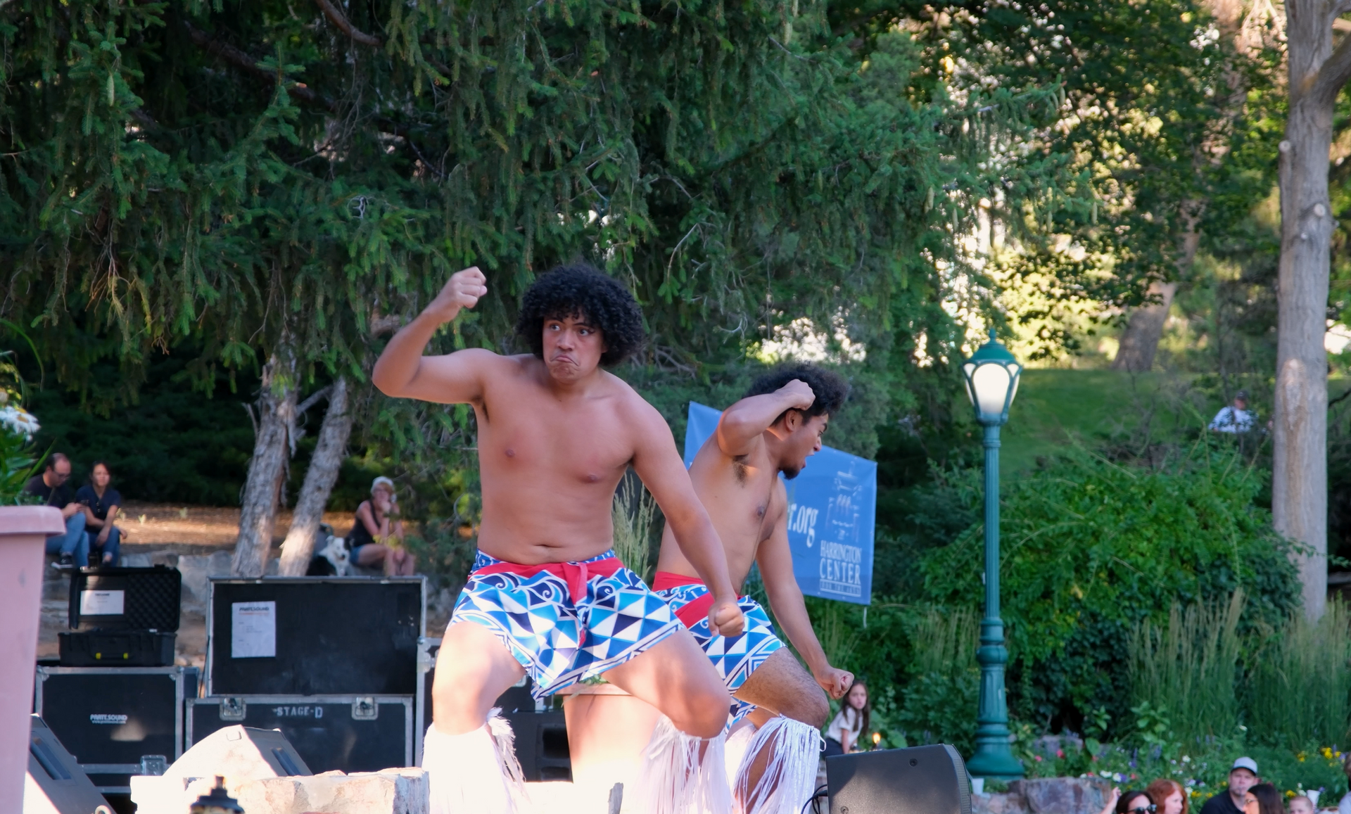 Two shirtless men are dancing in front of a crowd in a park.