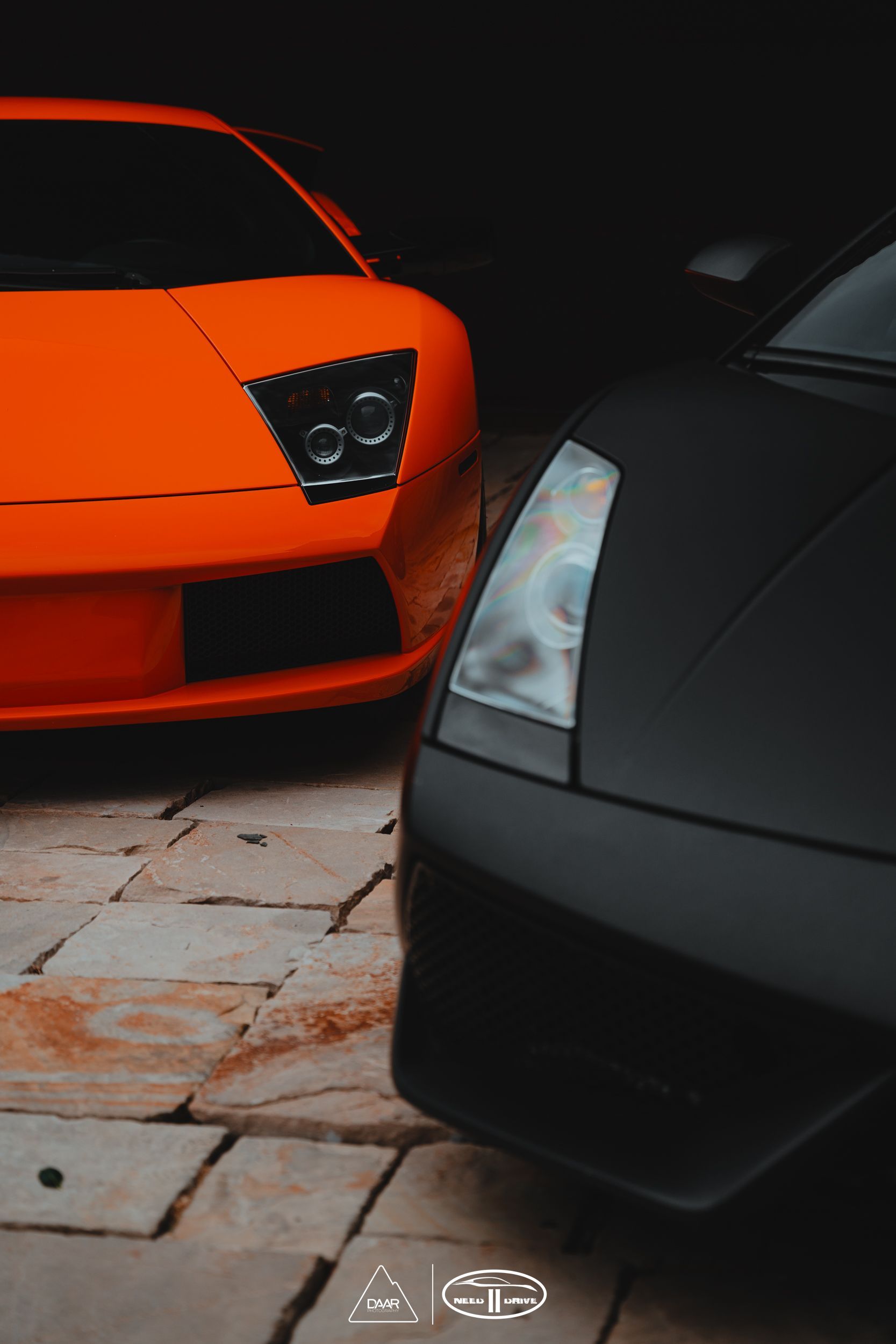 Orange Lamborghini Murcielago next to a black Lamborghini Gallardo in a stone garage.