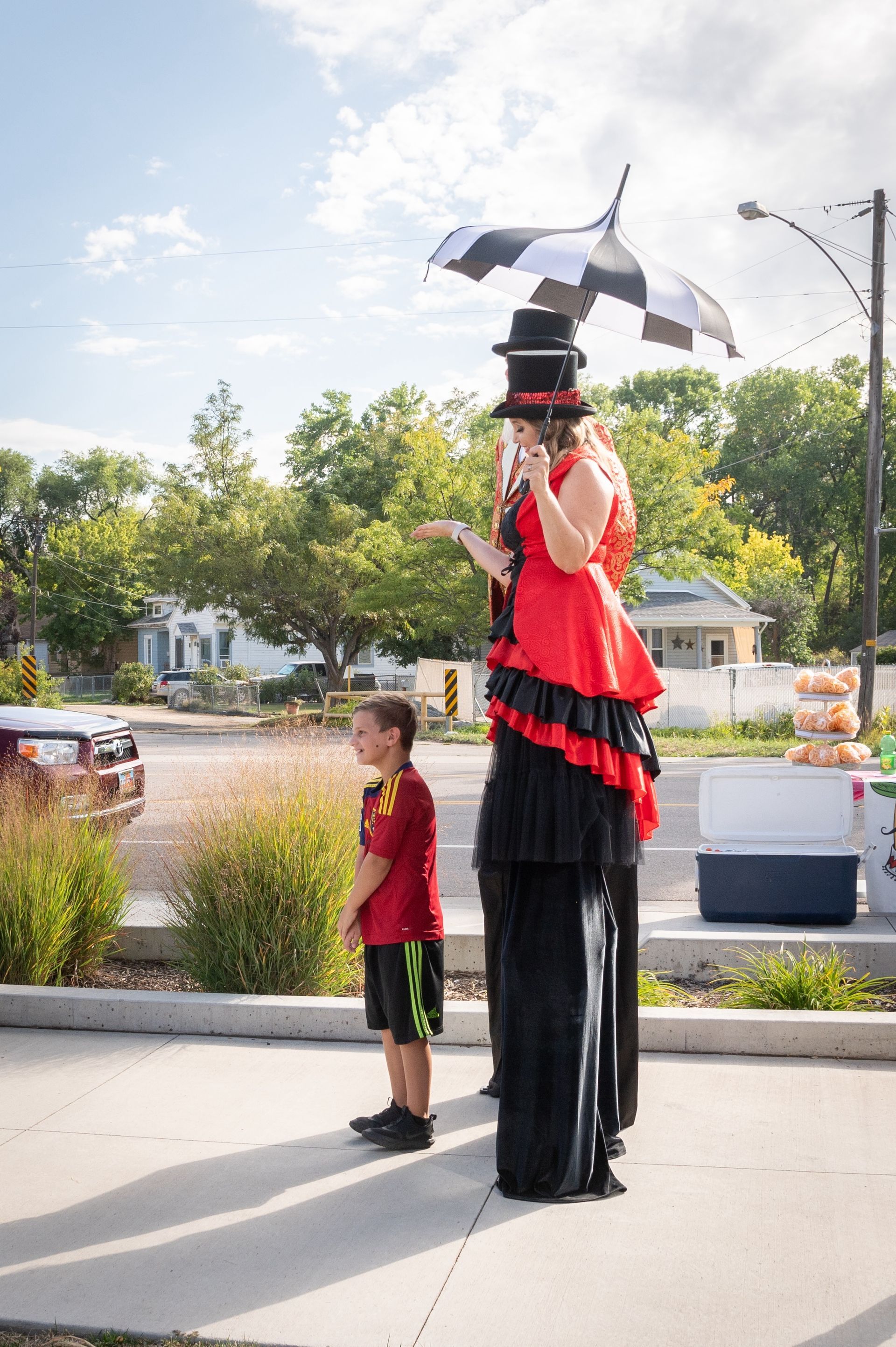 A woman on stilts with an umbrella on her head is standing next to a boy.