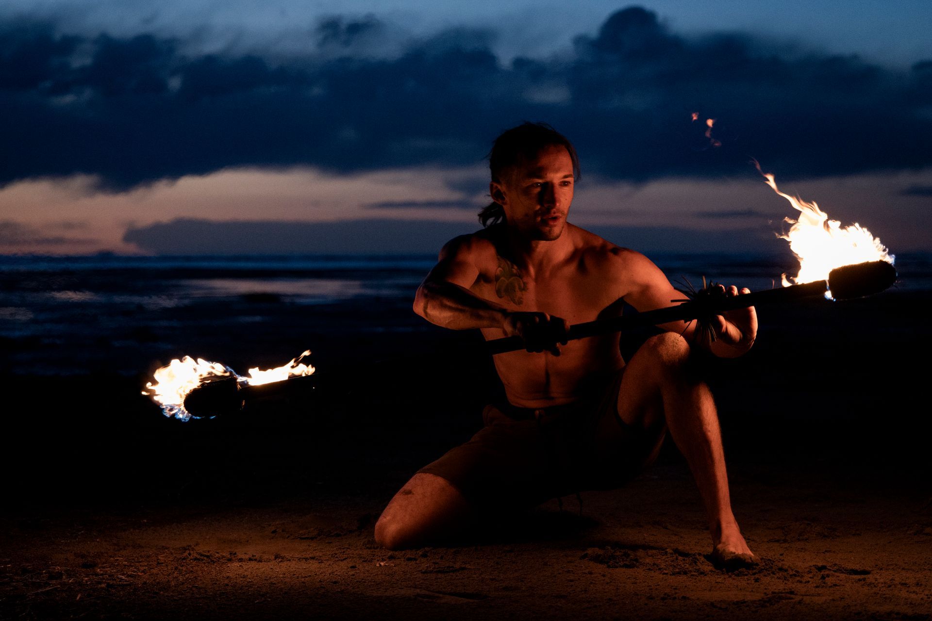 A man is kneeling down on the beach holding a fire torch.
