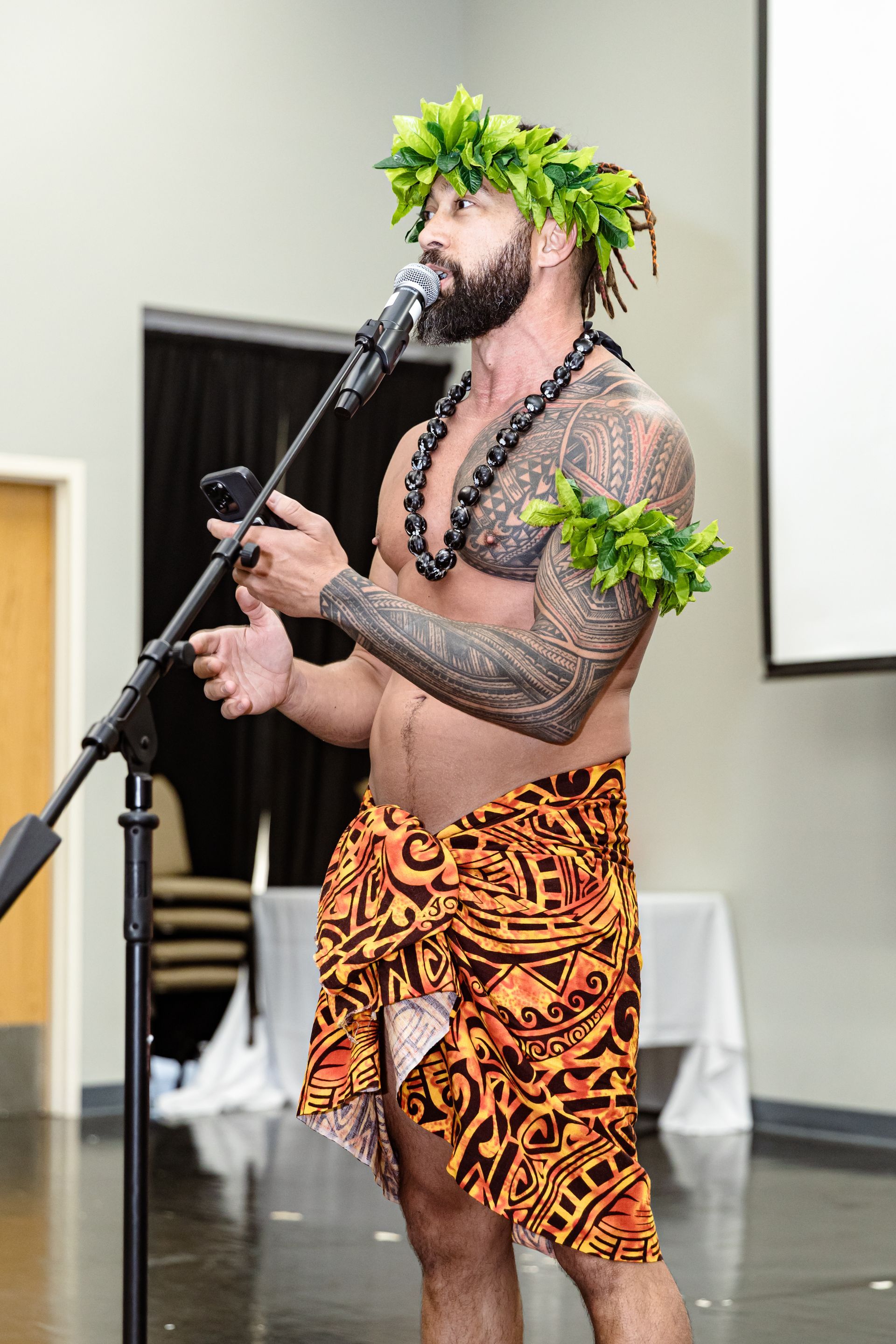 A shirtless man is singing into a microphone while wearing a lei.