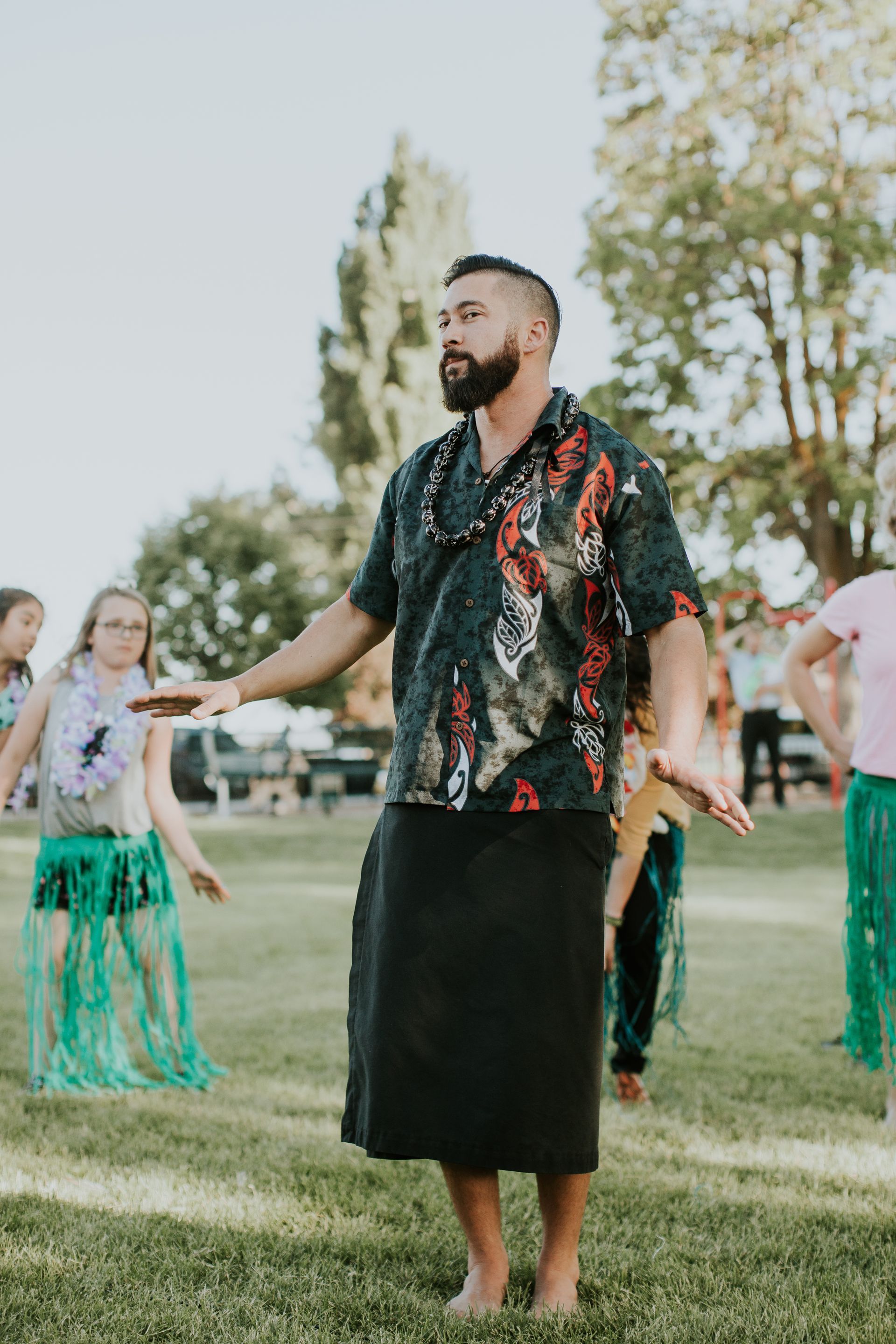 A man is standing in the grass with his arms outstretched in front of a group of children.