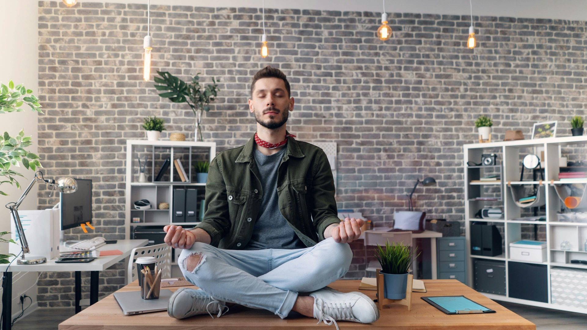 Man meditating cross-legged on a desk in an office. Brick wall background, neutral tones.