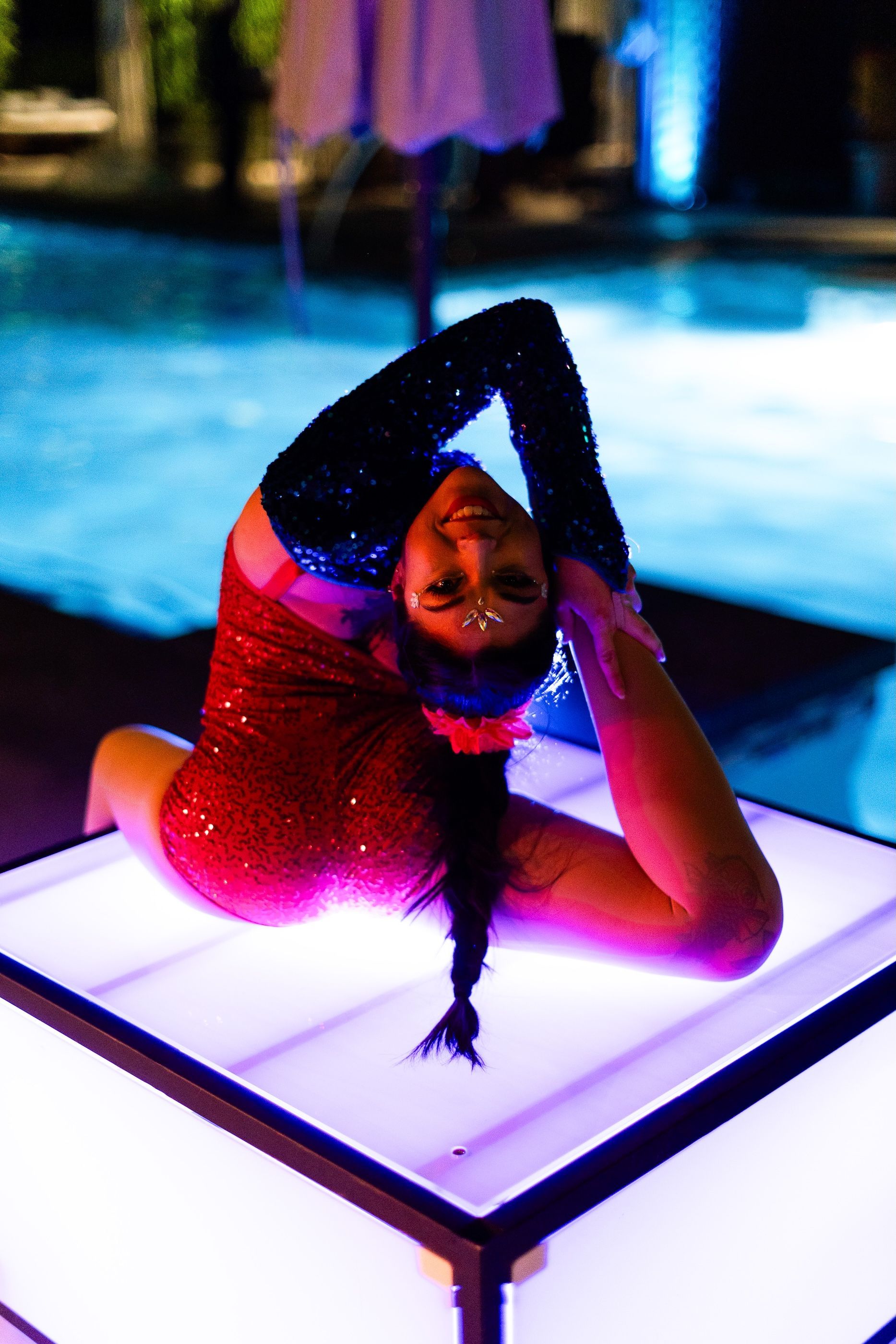A woman in a red bodysuit is doing a yoga pose on a table in front of a pool.