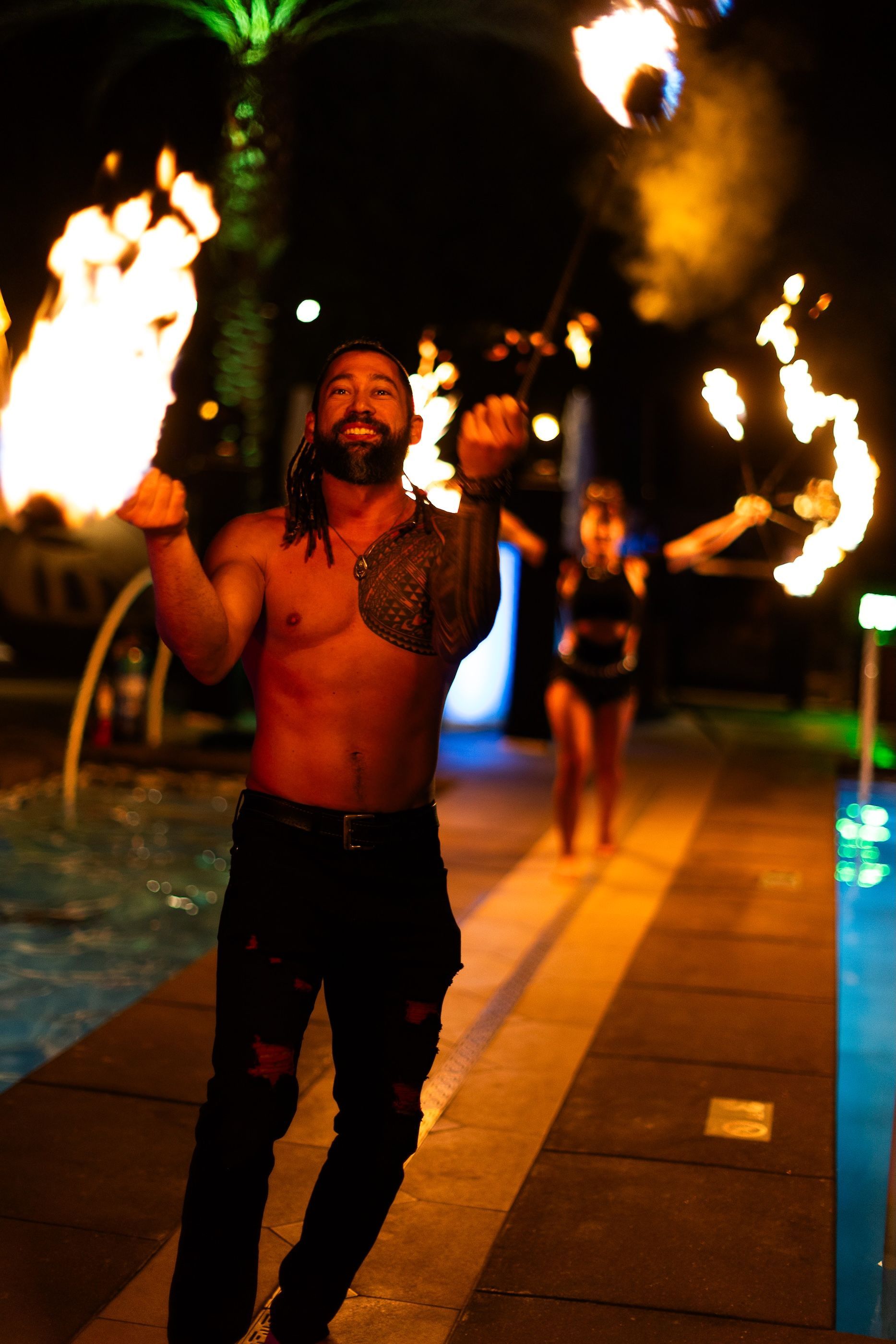 A man with a tattoo on his chest is holding a fire torch in front of a pool.