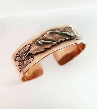A close up of a copper bracelet with leaves on it on a white surface.