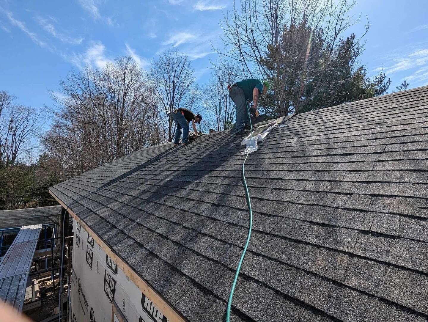 Two roofers on a gray shingled roof under a blue sky, working near trees.