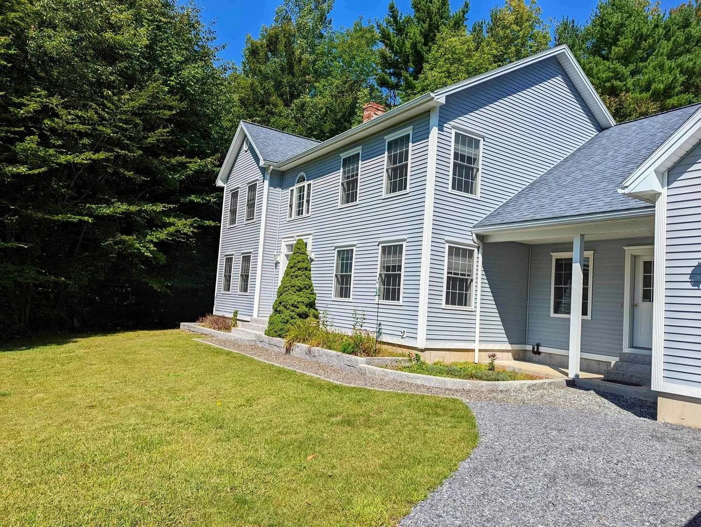 Two-story blue house with gravel driveway and green lawn, surrounded by trees on a sunny day.