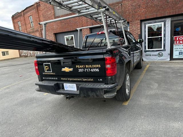 Black pickup truck with ladder rack, parked outside a brick building. The truck bed holds dark material.