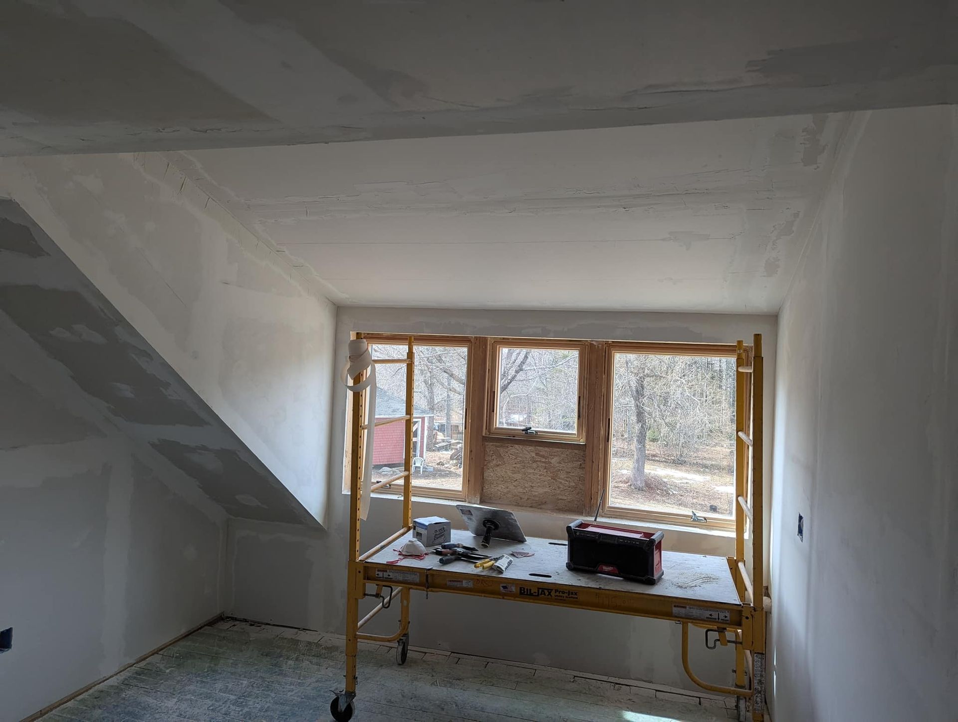 Interior room under construction, featuring a window and scaffolding. Walls are white, a light-colored scaffold, and a view of trees outside.