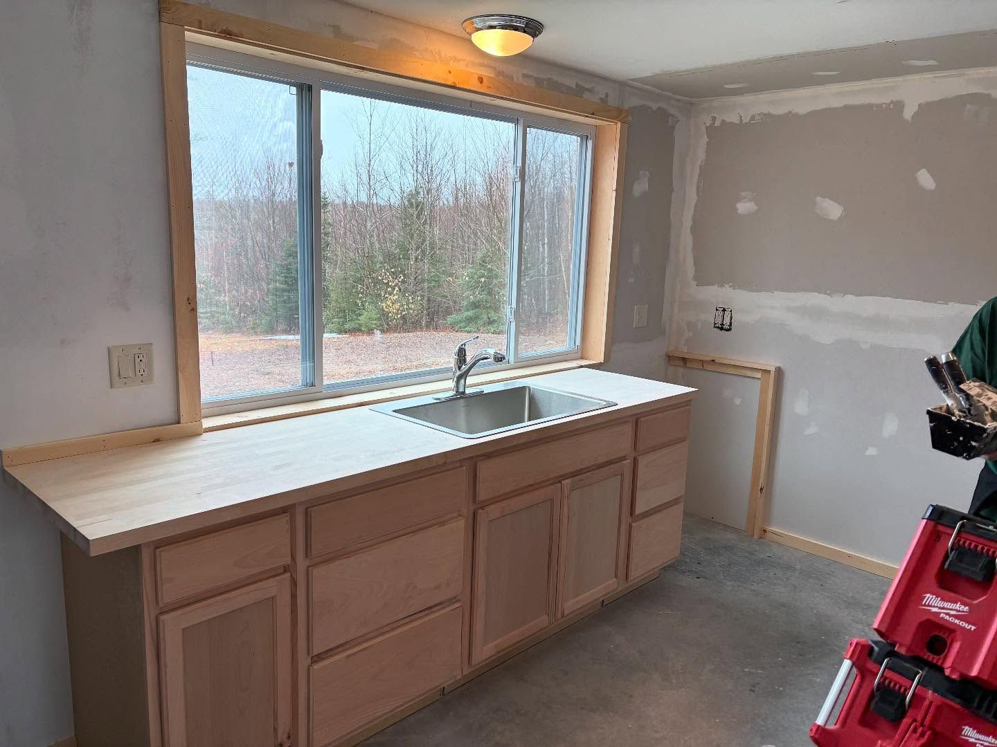 Kitchen under construction, with unfinished cabinets, sink beneath a window, and drywall.