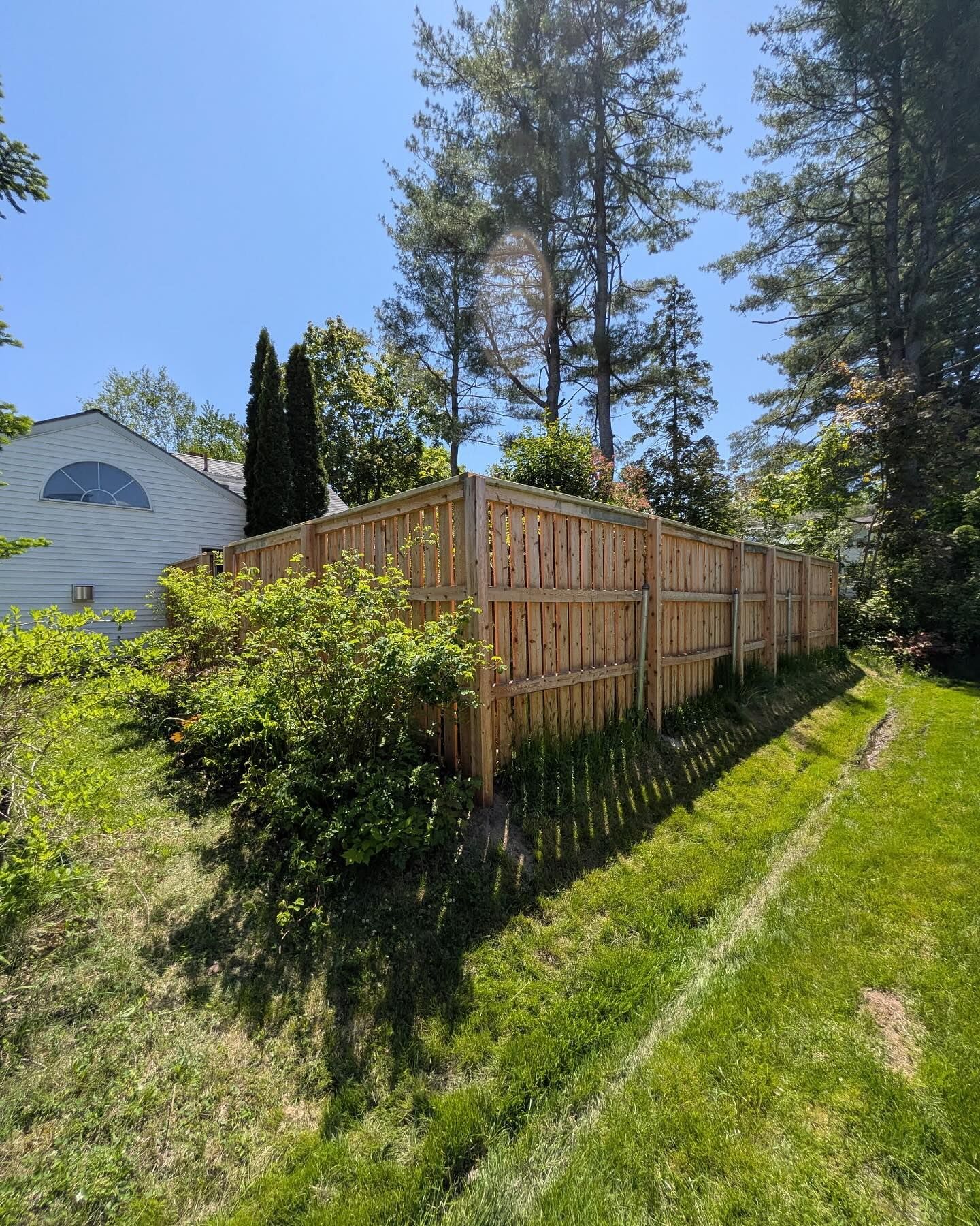 Wooden fence bordering a grassy yard on a sunny day, with trees and a house in the background.