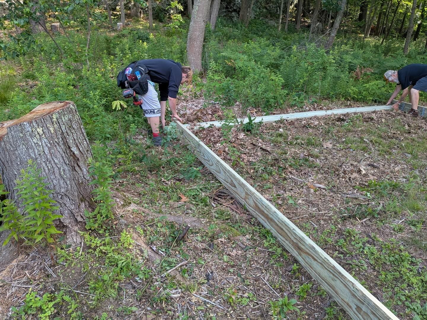 Three people working on a wooden fence outdoors in a wooded area.
