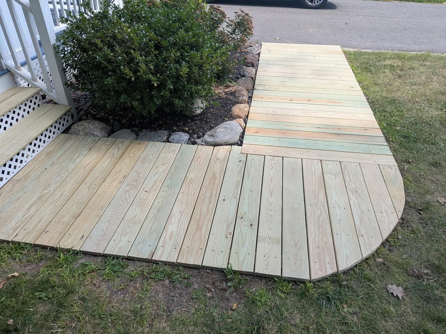Wooden ramp and walkway leading to a house entrance, beside a bush and grass.