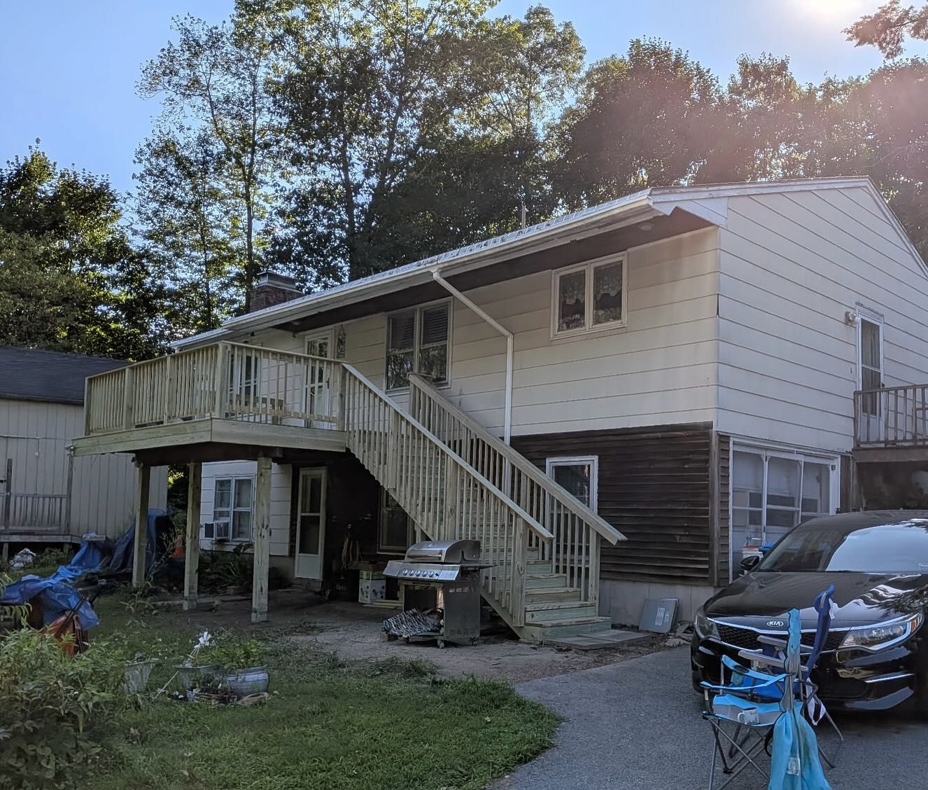 Two-story house with wooden deck, stairs, and parked car. House has white siding, surrounded by trees.