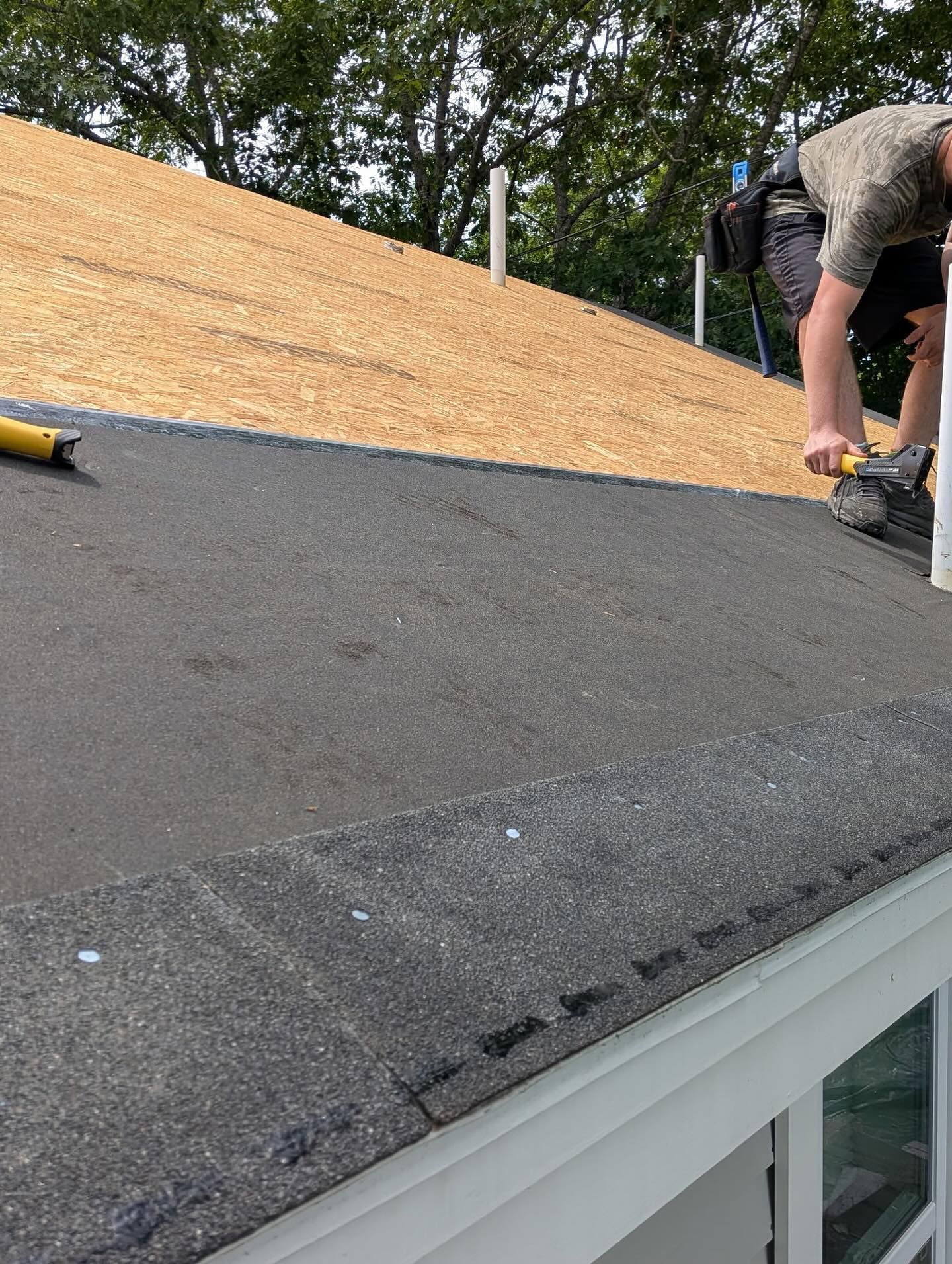 Roofer working on a dark asphalt shingle roof, with a new wood shingle layer behind, sunny day.