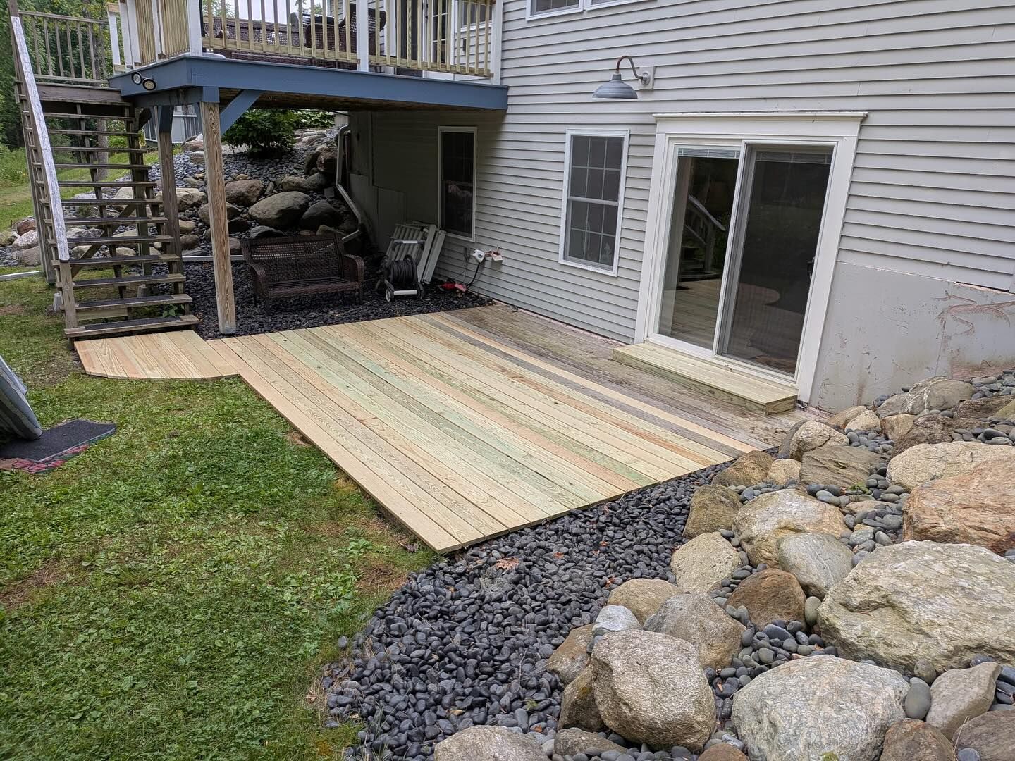 Wooden deck and patio area below a house, with stone landscaping and grass.