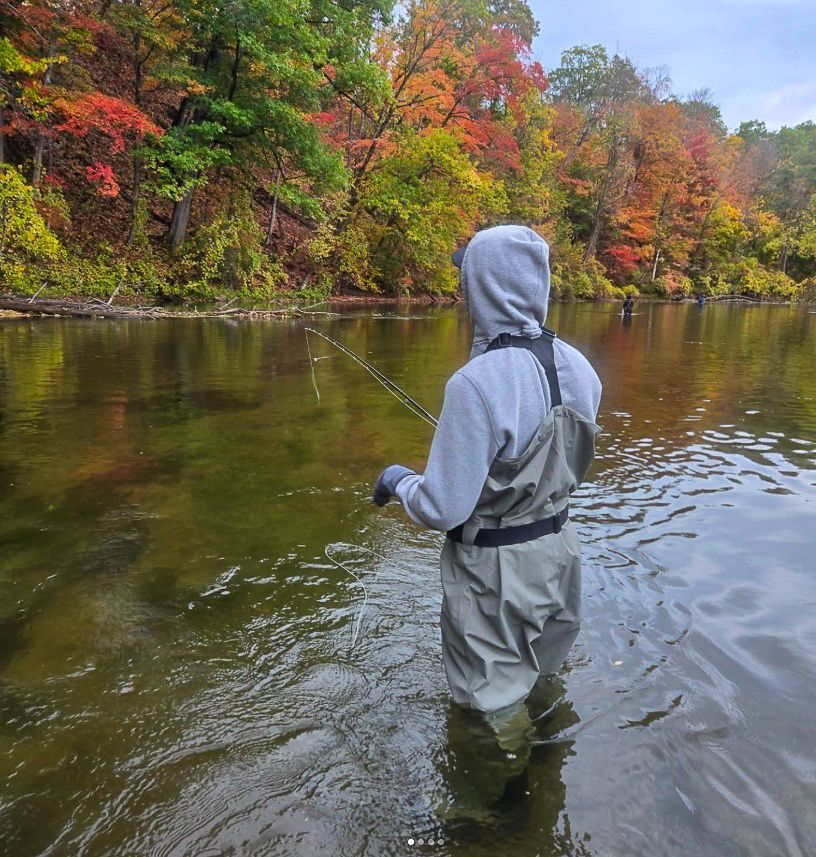 A person wearing a grey hoodie and waders fly-fishes in a river surrounded by vibrant autumn trees.