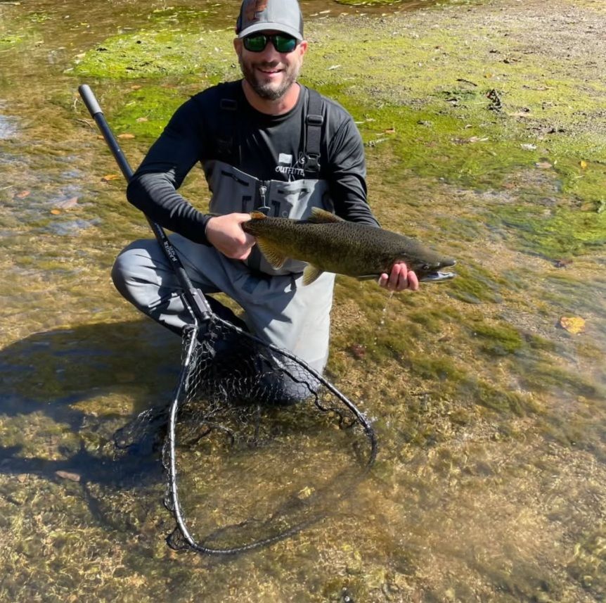 A person wearing a cap, sunglasses, and waders kneels in a shallow stream, smiling while holding a freshly caught fish.