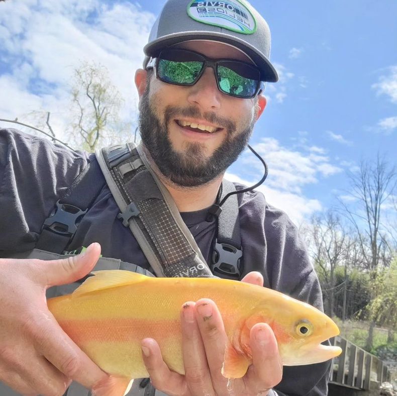 David Kuhn, Founder of The Rods for Wheels Organization,  smiling while holding a bright yellow-orange trout outdoors against a blue sky.