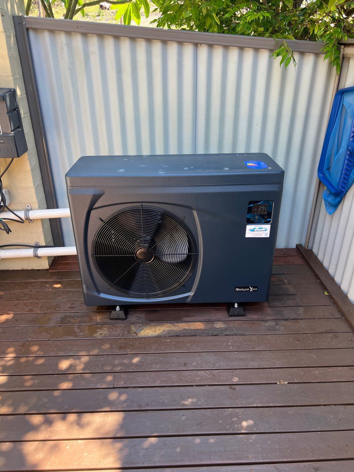 Outdoor air conditioning unit beside a corrugated metal fence and wooden deck, with a blue hose nearby